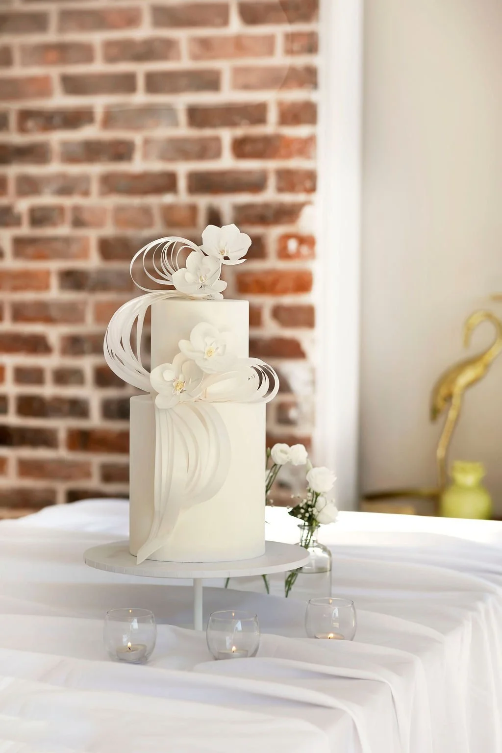 Elegant white wedding cake with floral decoration on a white table, with a brick wall in the background.