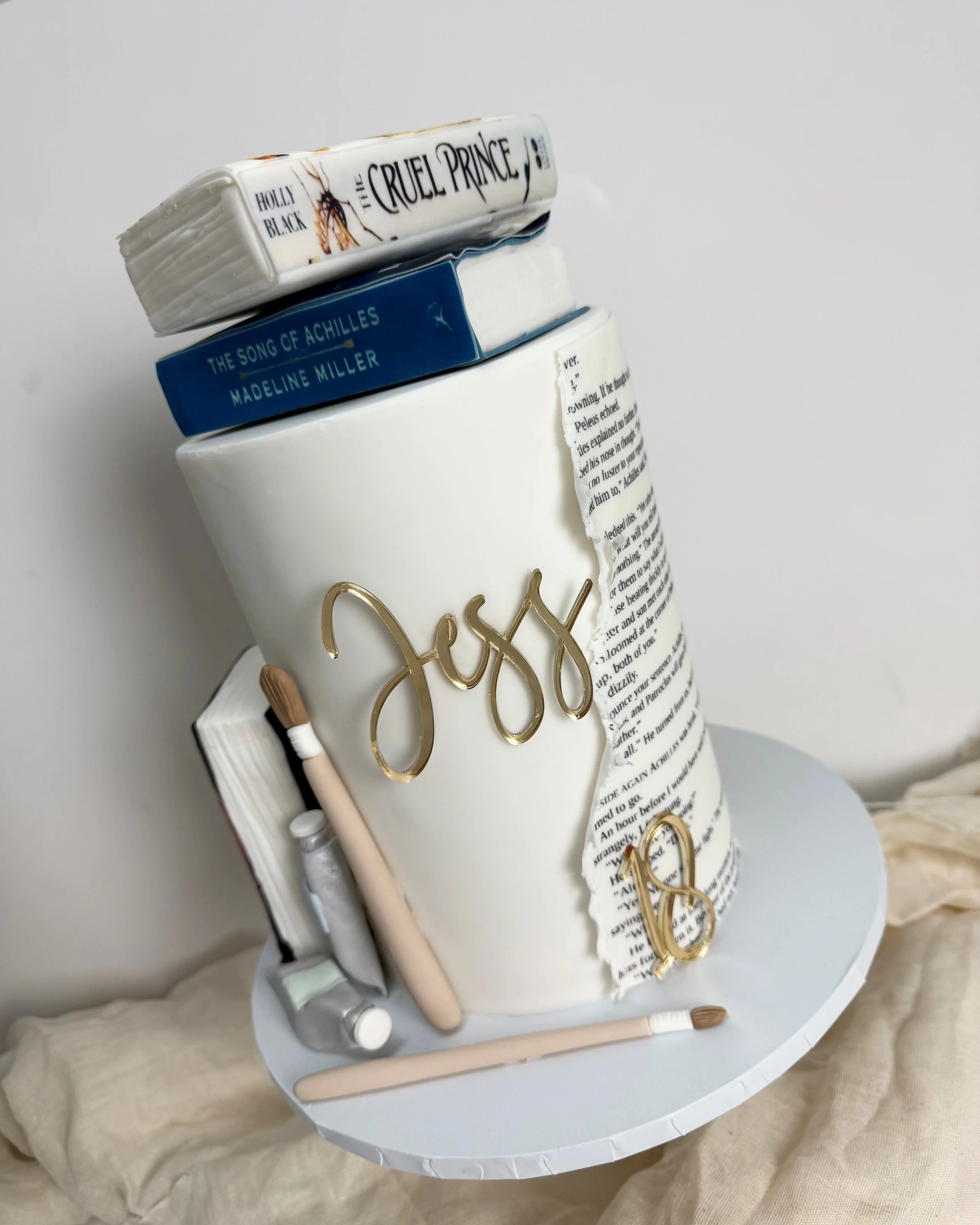 A baby shower cake decorated as a white disposable cup with the word 'Joy' in gold and the number 8. There are books, makeup brushes, and other items on top and around the cake.