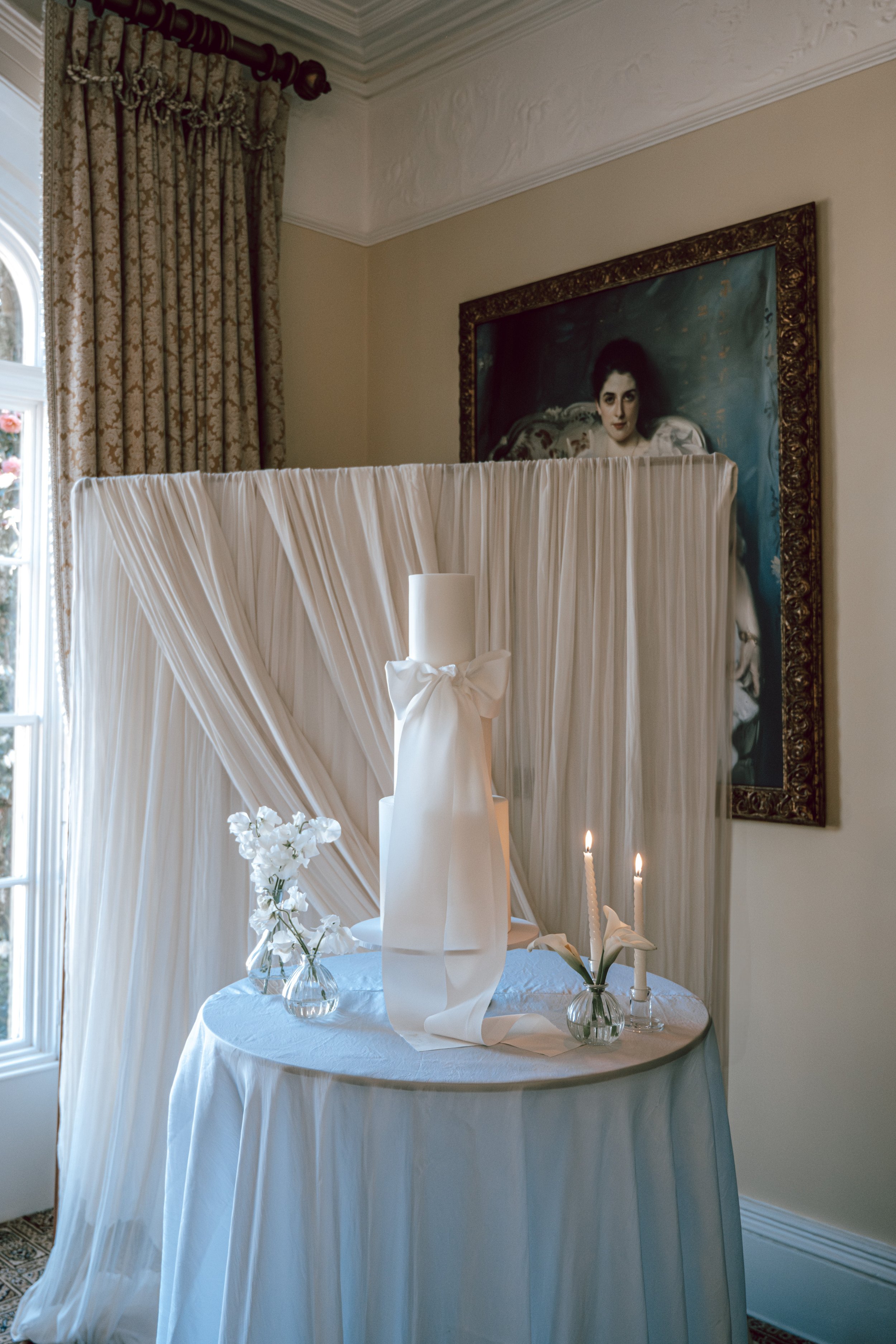 A round table draped with white cloth, decorated with white flowers in small vases, two lit candles, and a large white candle with a bow, set indoors near a window with patterned curtains, with an ornate gold-framed portrait of a woman hanging on the wall behind.