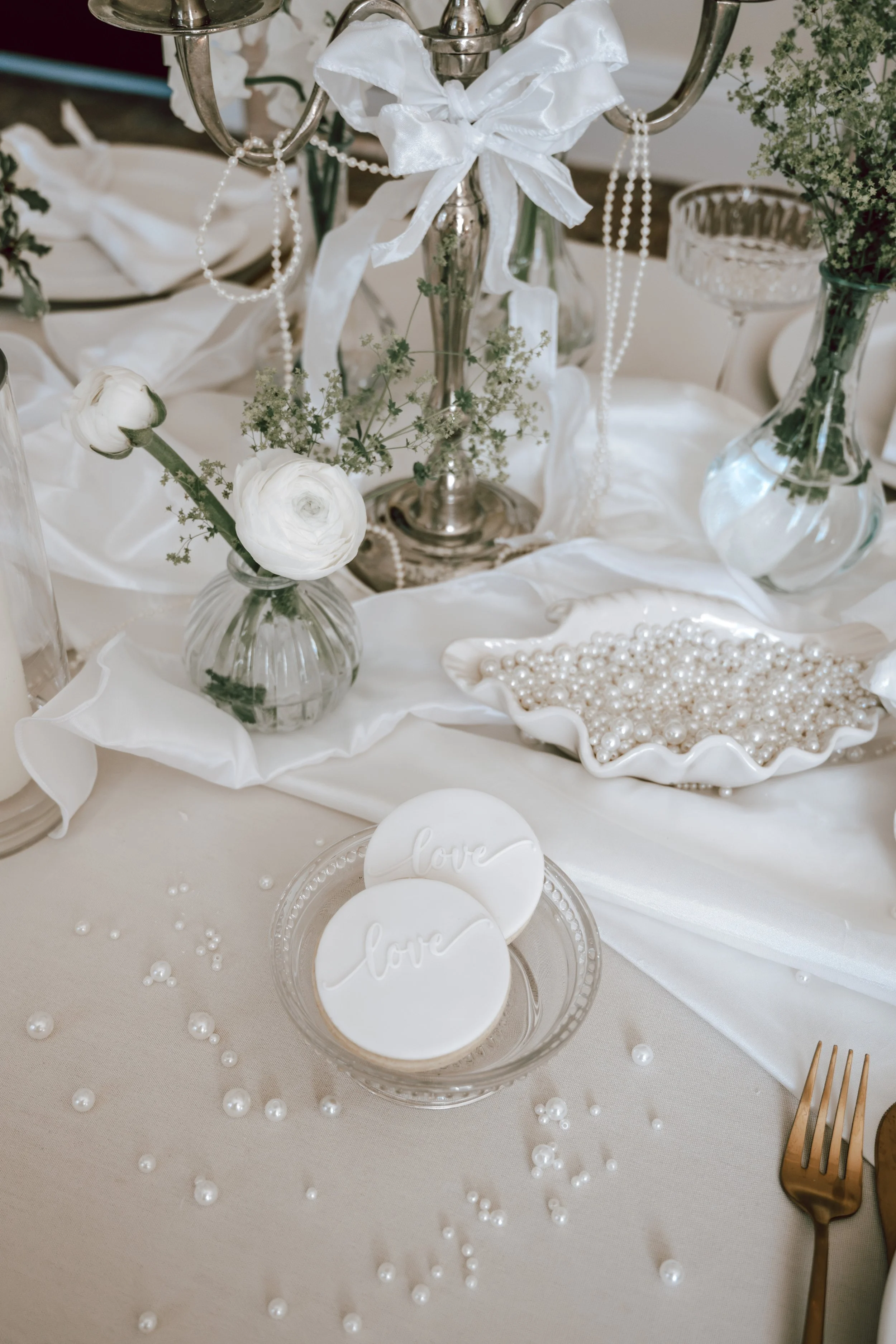 Elegant table setting with white and silver decorations, including flowers, pearls, and love-themed cookies.