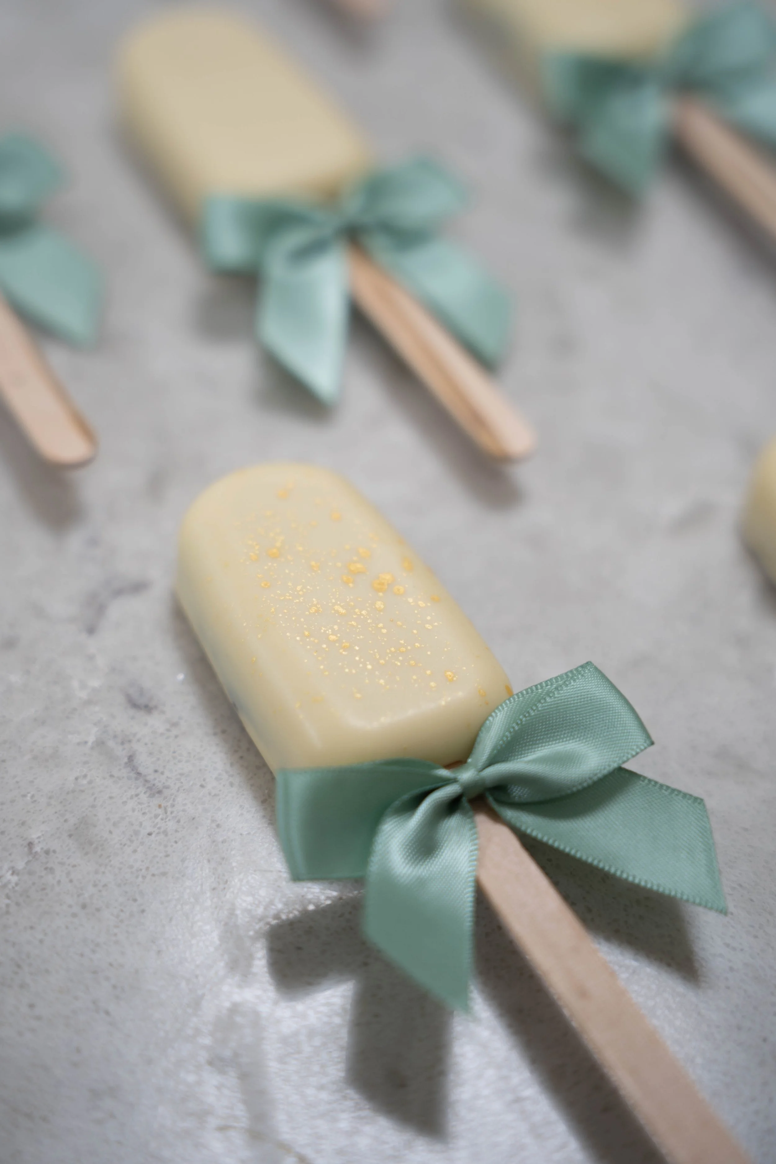 Cream-colored popsicle with yellow speckles and a green satin ribbon tied in a bow on a gray surface.