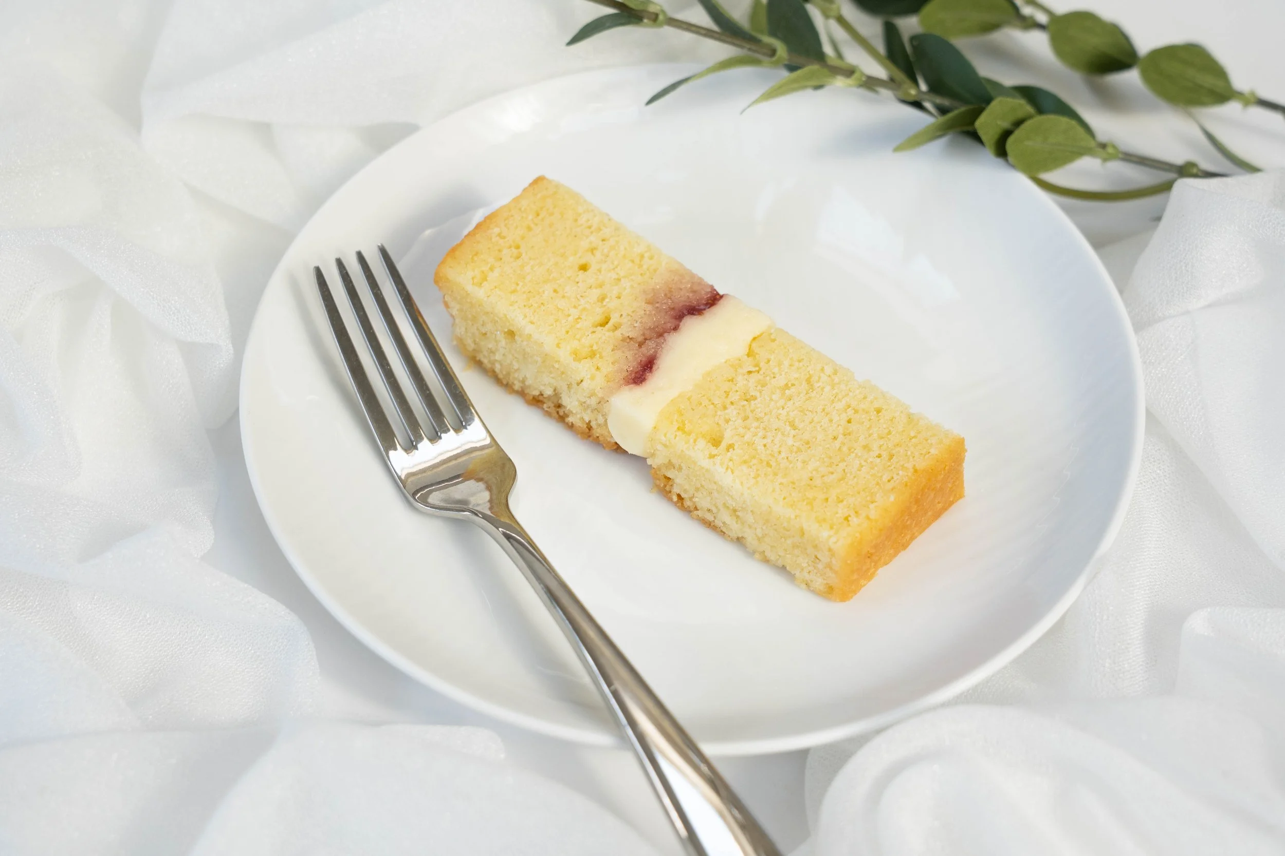 A slice of cake with cream and berry filling on a white plate, accompanied by a fork, on a white tablecloth with green leaves in the background.