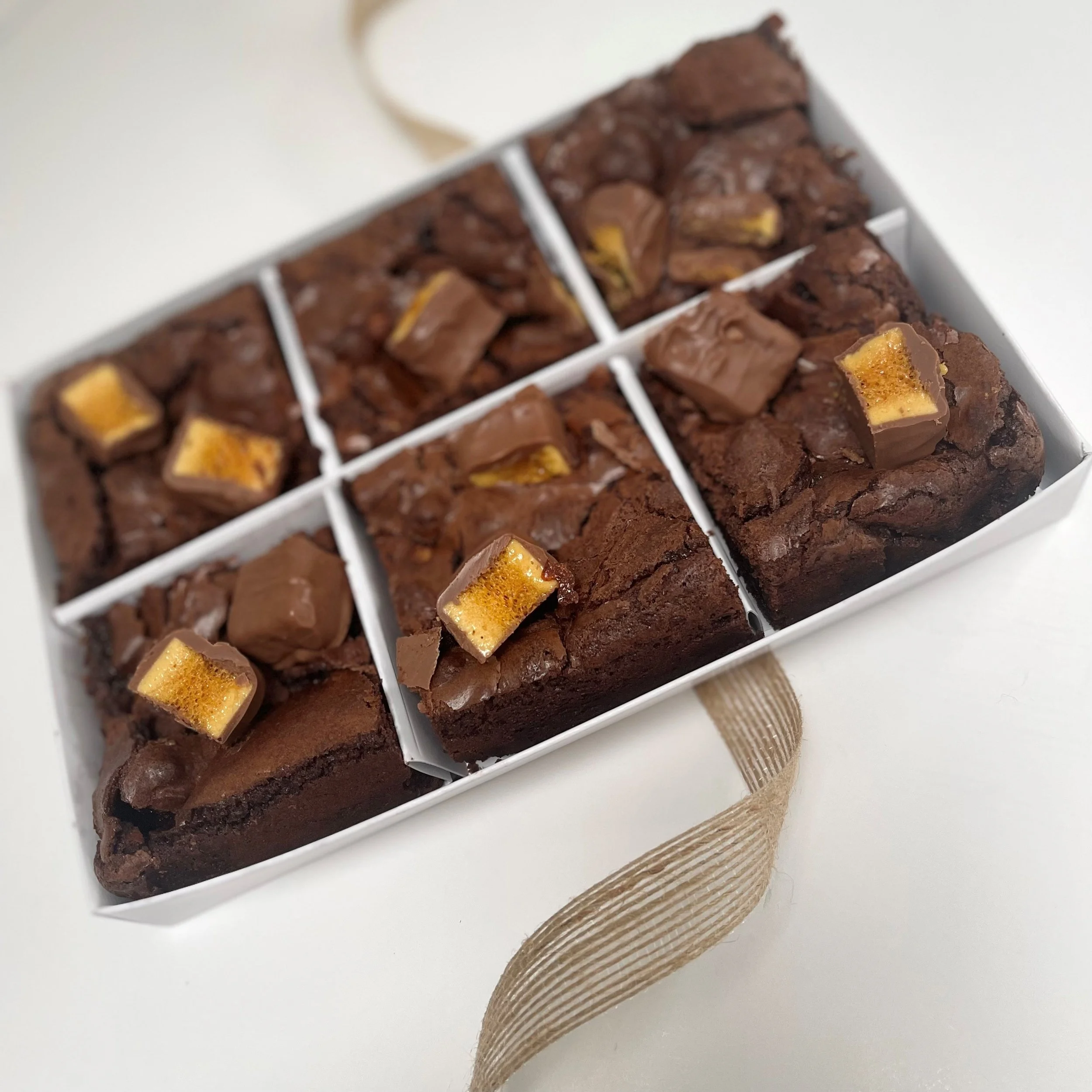 Assorted brownies topped with caramel-filled chocolate candies in a white box with a beige ribbon handle.