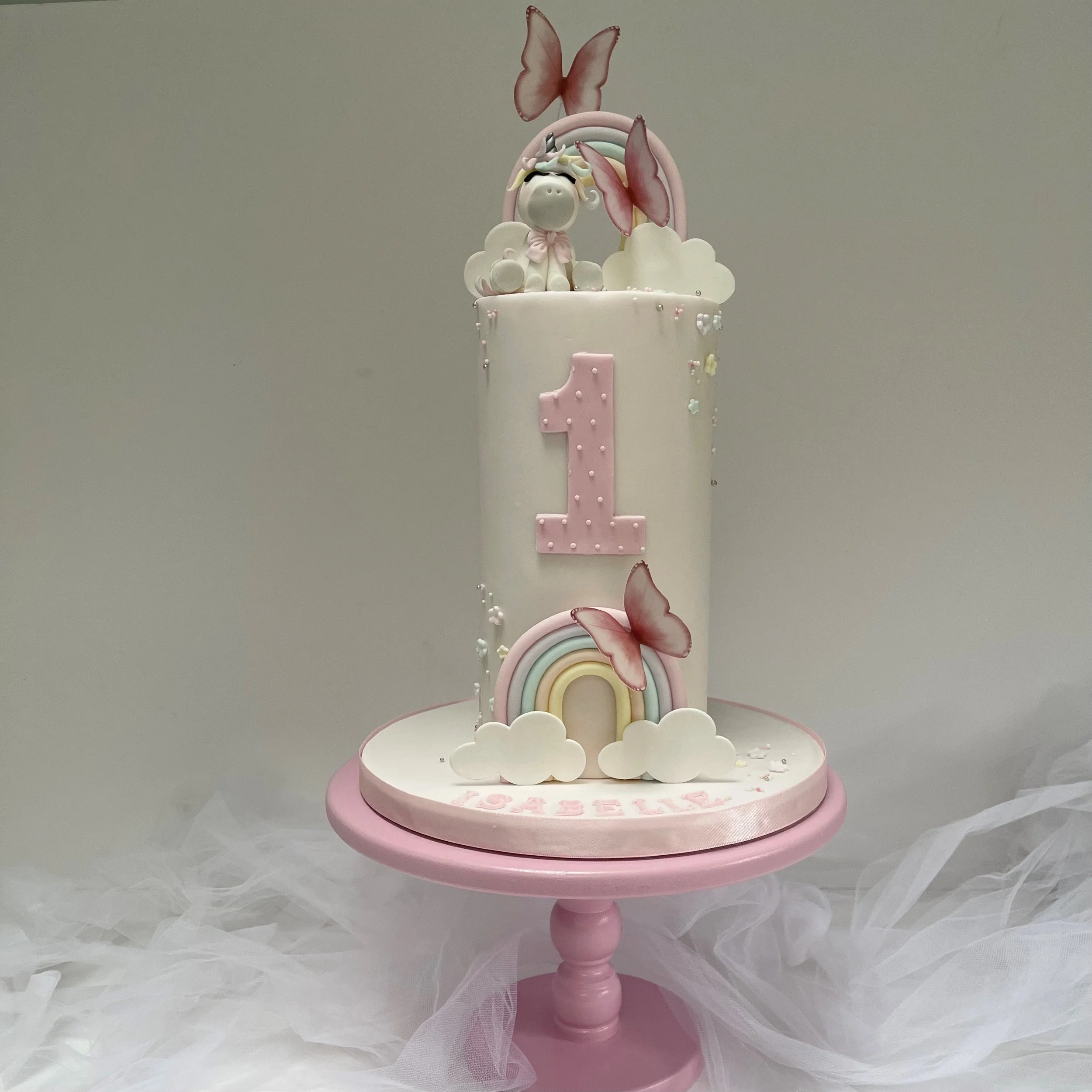 A unicorn-themed first birthday cake with a pink and white color scheme, featuring a unicorn figurine, rainbow, clouds, pink butterfly decorations, and a large pink number one on the front.