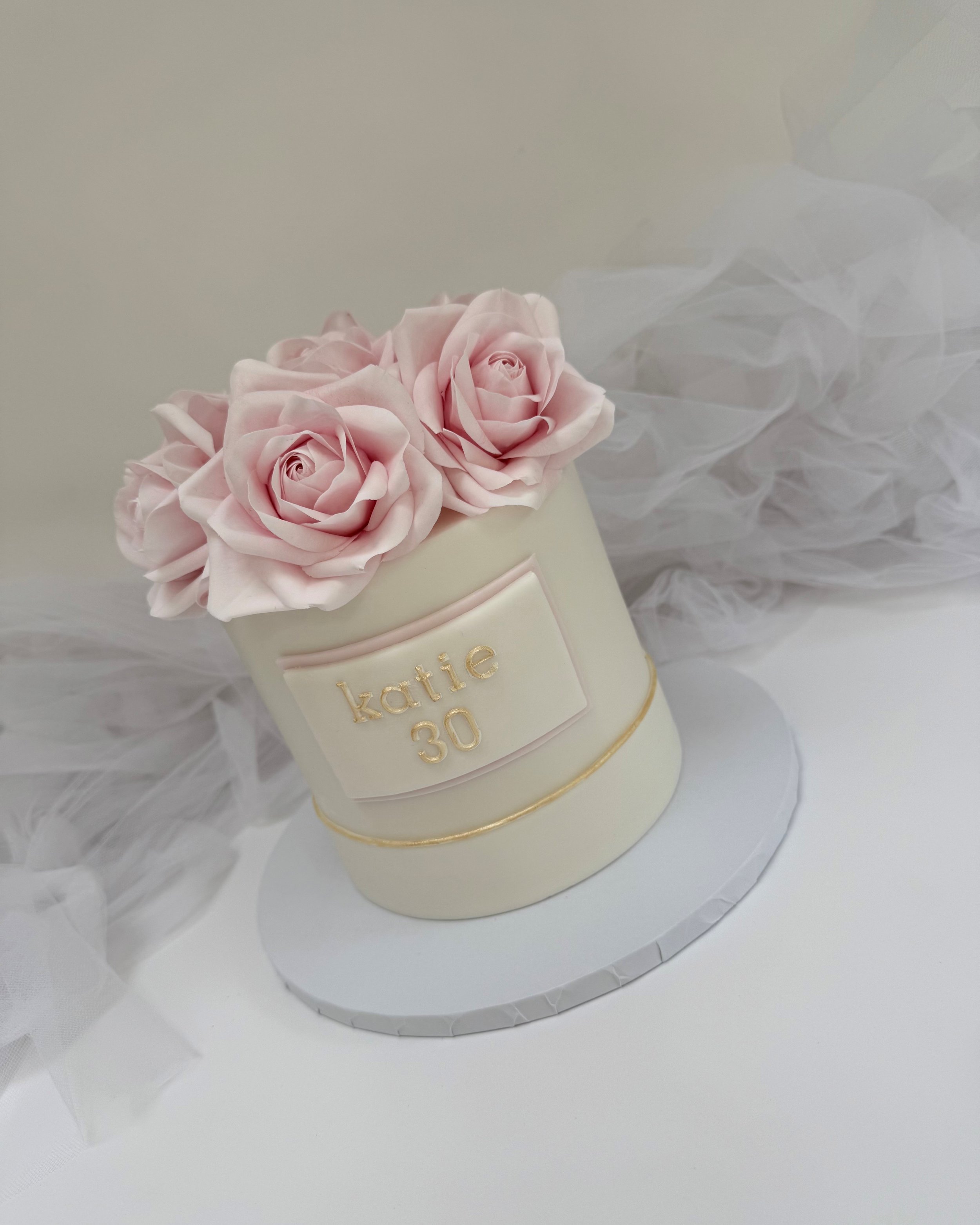A white cylindrical cake with pink roses on top, personalized with gold lettering that says 'katie 30,' sitting on a round white base with a gold line, surrounded by white tulle fabric.