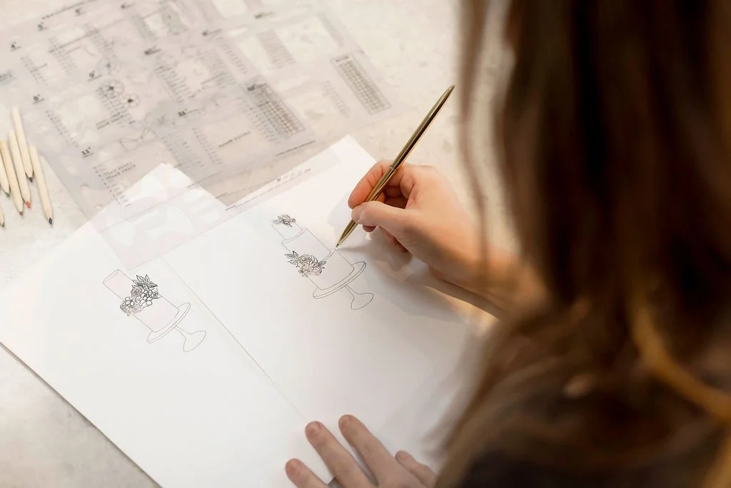 Rebecca is designing a cake on a sheet of paper, with colored pencils and printed diagrams nearby.