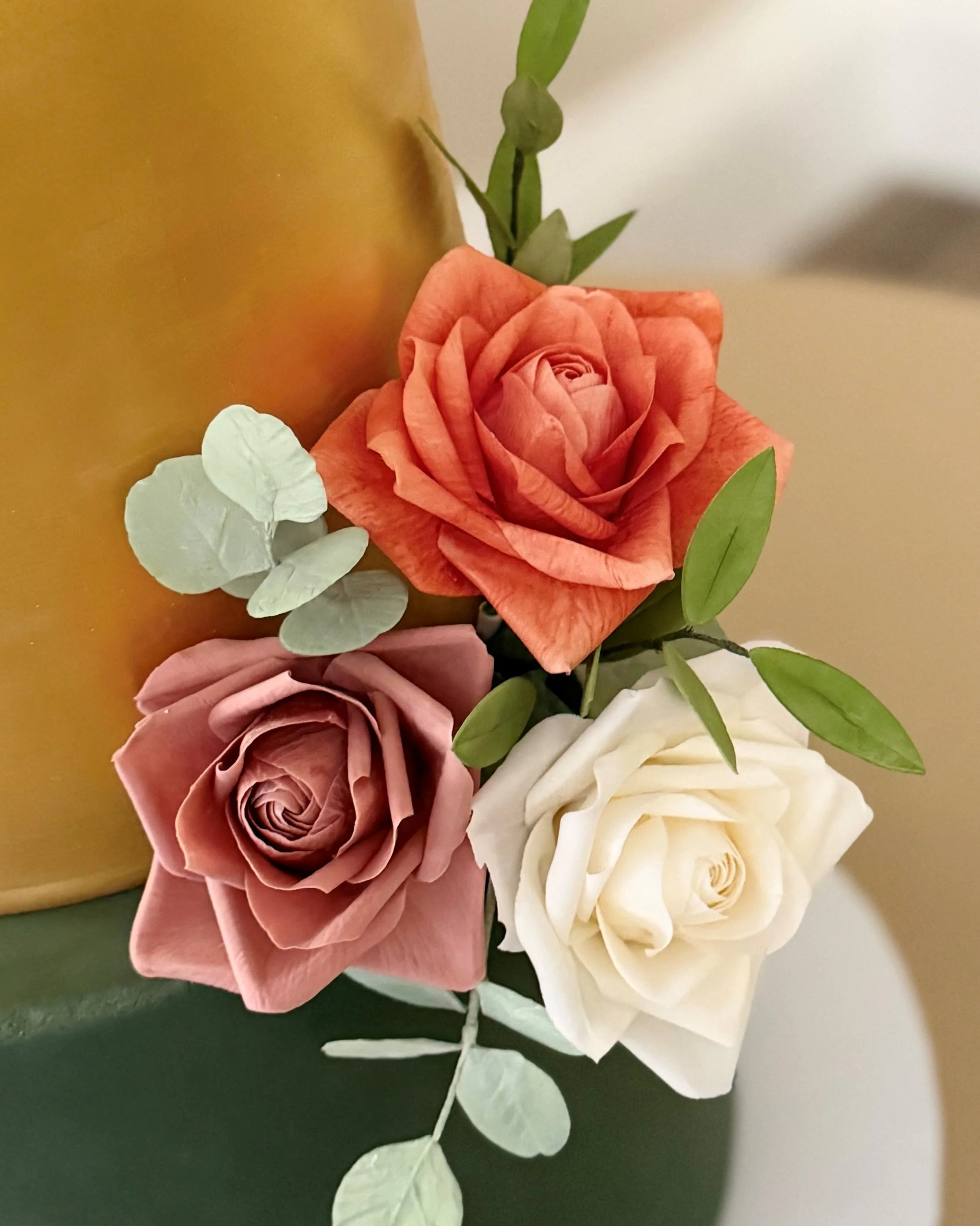 A bouquet of three roses in pink, orange, and white, accompanied by green leaves and eucalyptus, placed against a blurred background.
