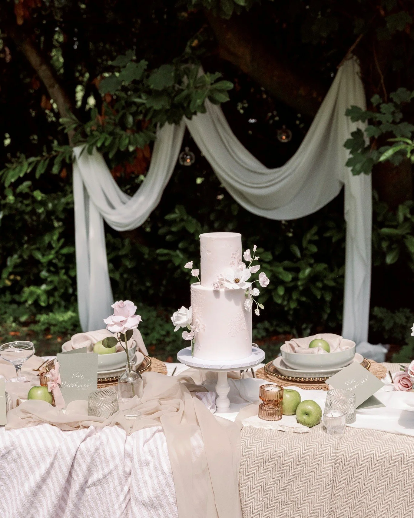 Elegant wedding dessert table with a two-tier white cake decorated with white flowers, set outdoors with greenery, draped white fabric, and hanging lights in the background.