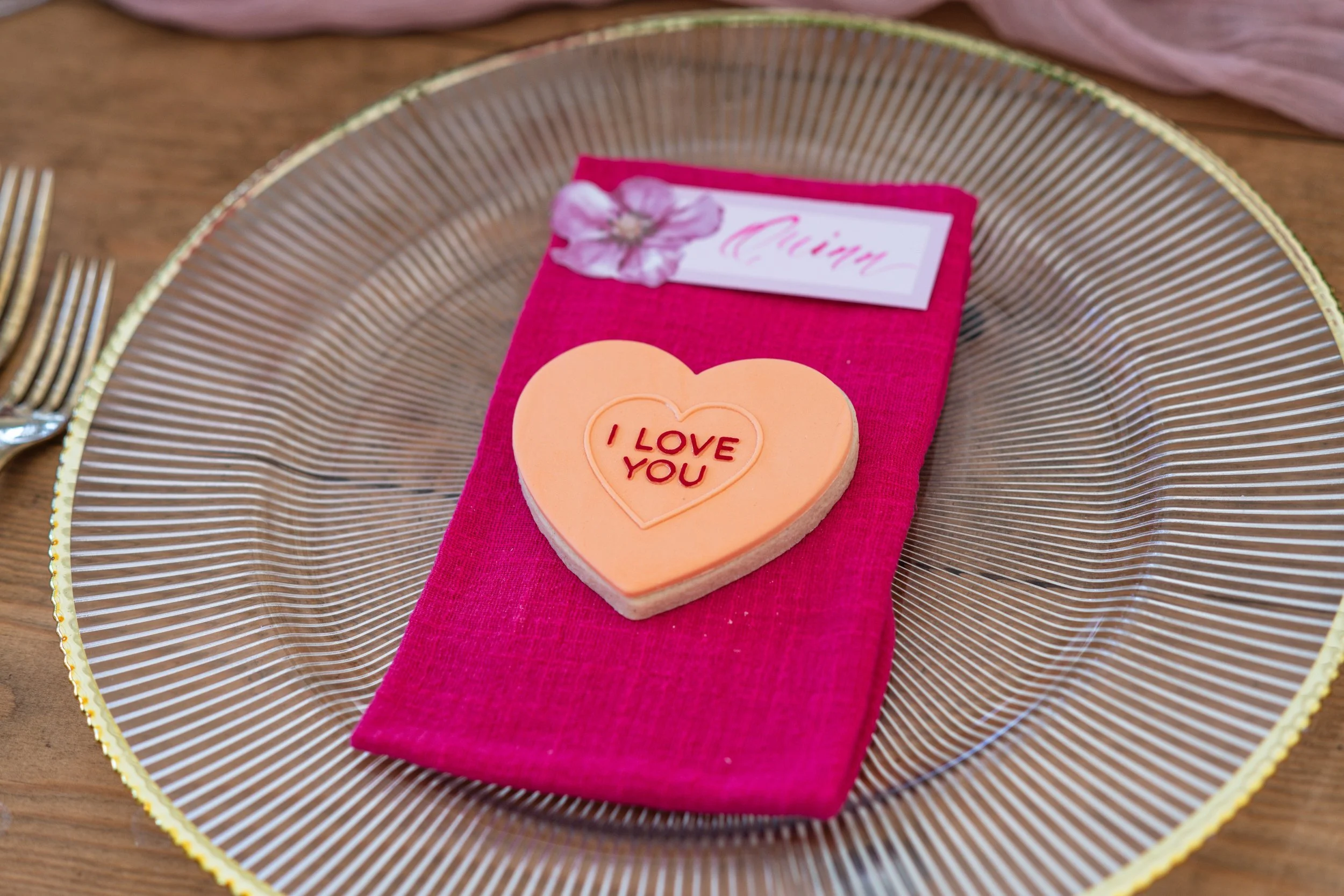 A pink napkin with a heart-shaped cookie that says 'I LOVE YOU', a white name tag with pink writing and a flower, all placed on a clear glass plate with a gold rim, set on a wooden table with silver forks on the sides.