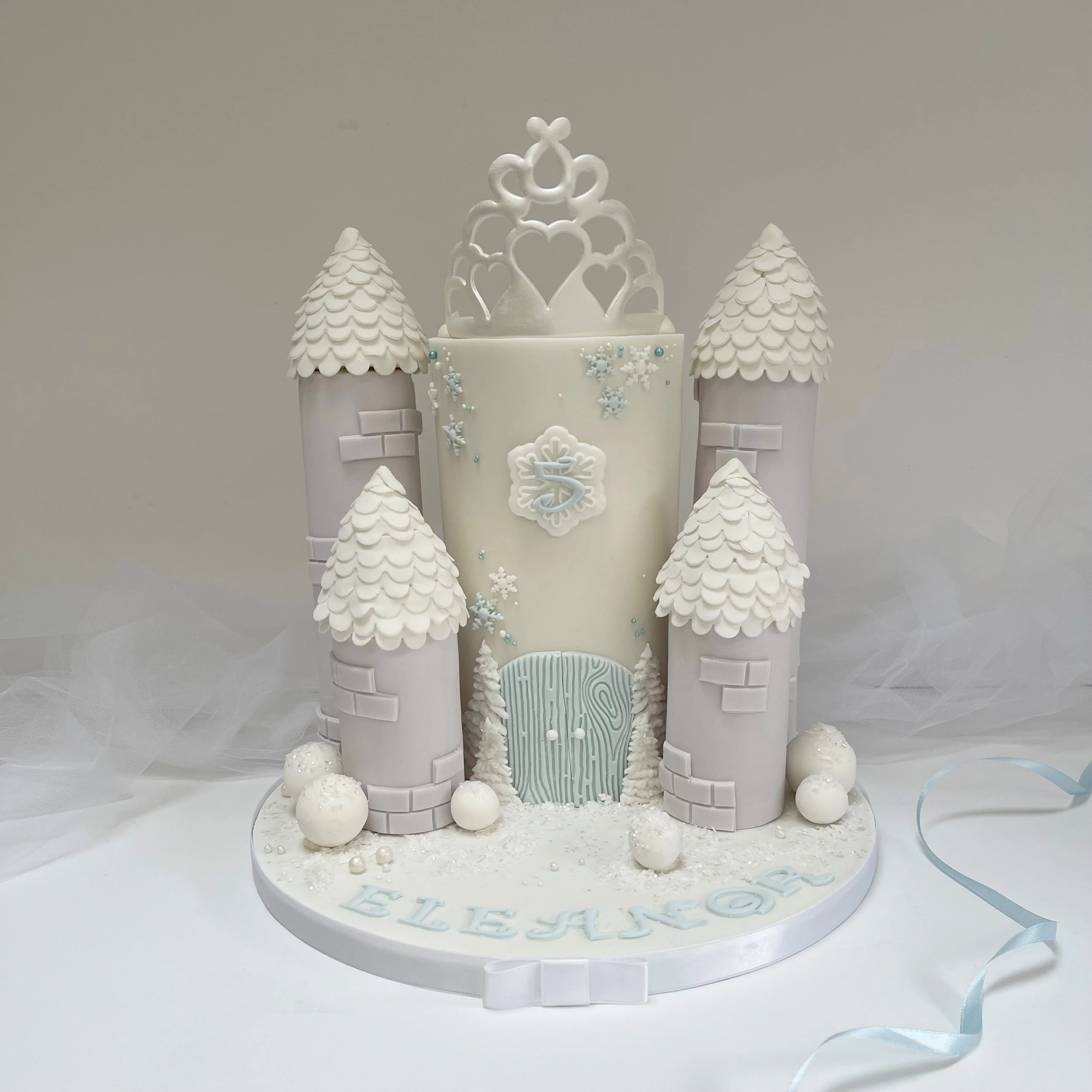 A white castle-themed cake with snowflakes, a wooden door, and a princess crown topper, decorated in icy blue and white colors.