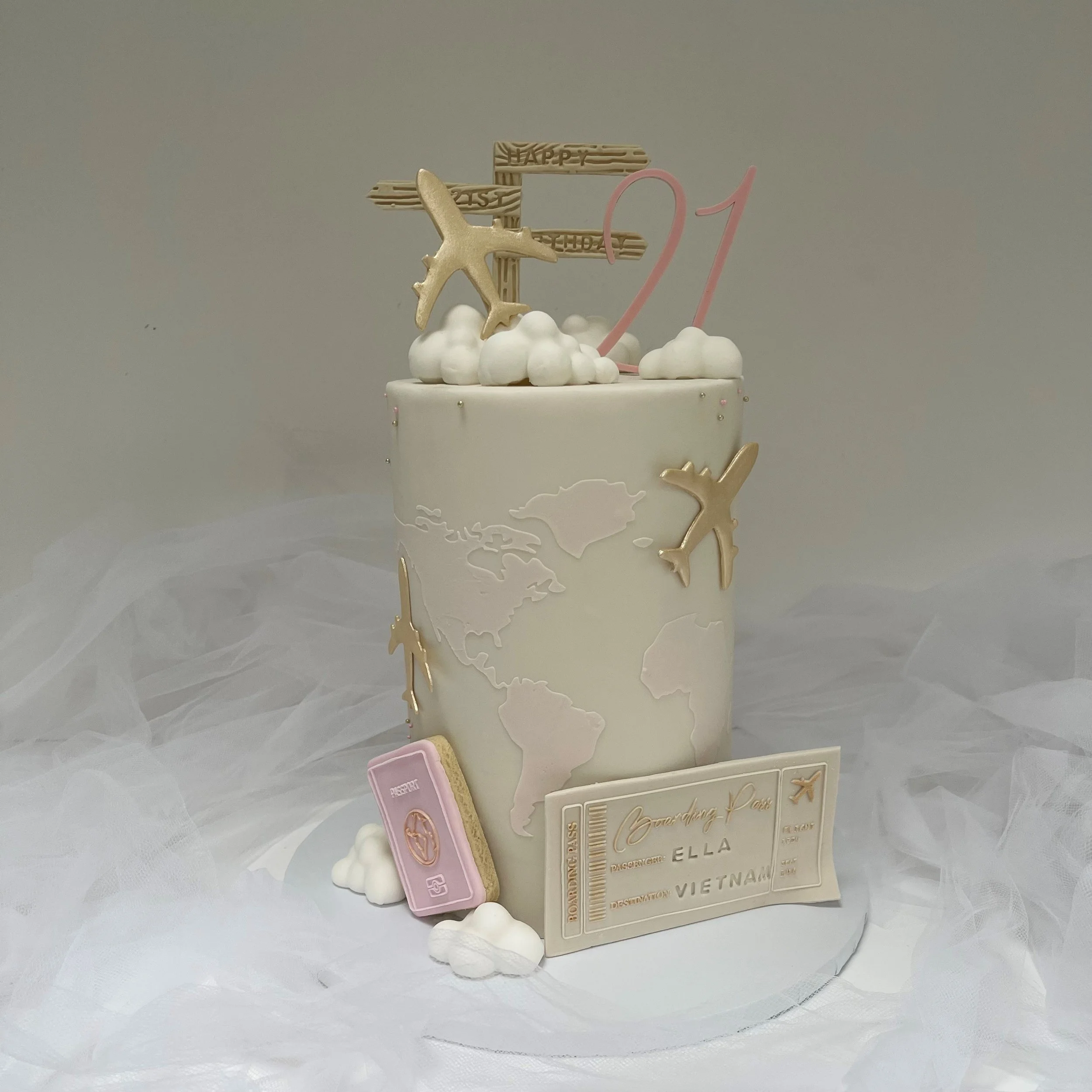 A white birthday cake decorated with gold airplane charms, a pink number 21, and a world map design, surrounded by clouds and travel-themed decorations.
