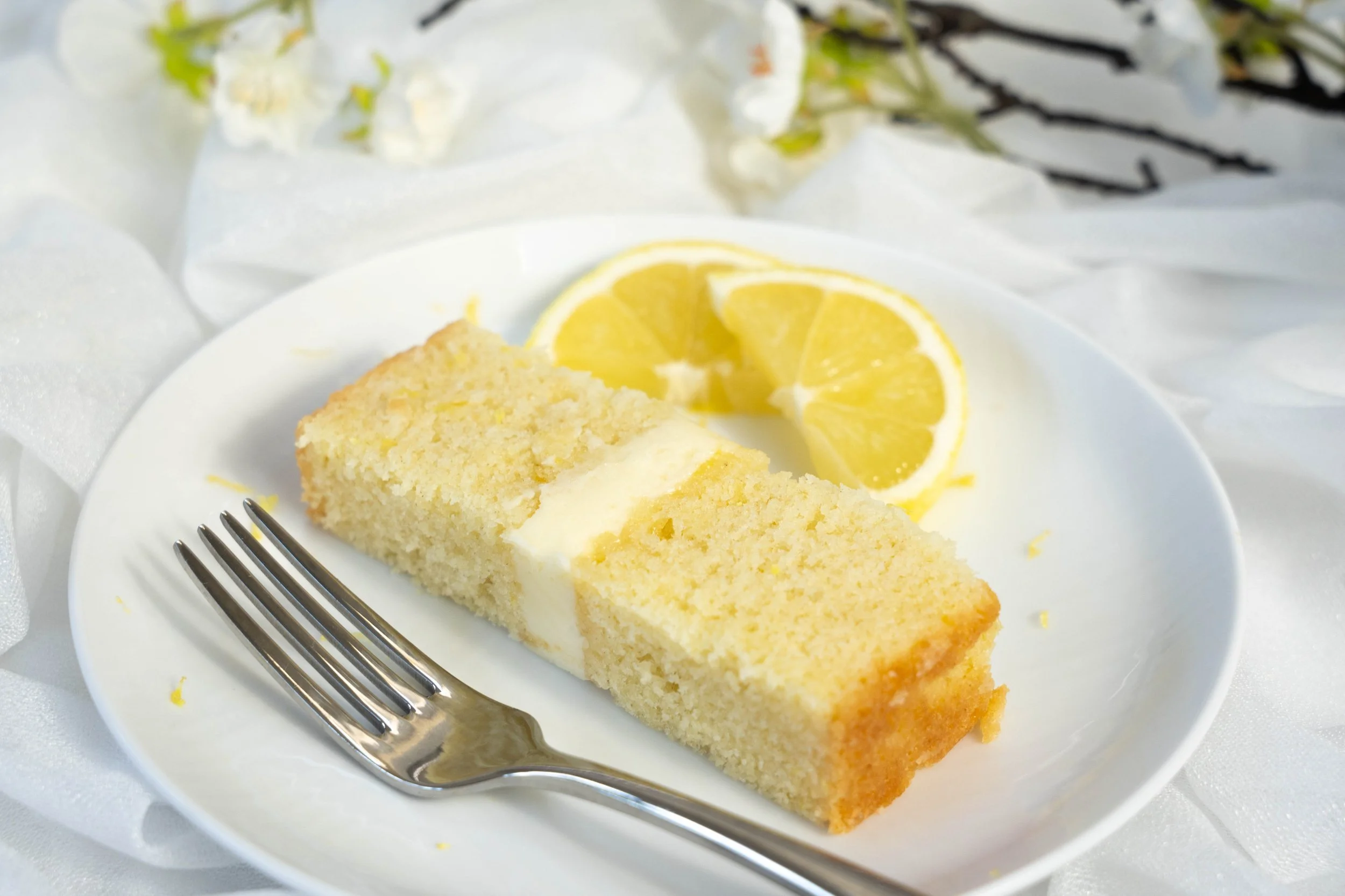 A slice of lemon cake on a white plate with lemon slices and a fork, surrounded by white decorative fabrics.