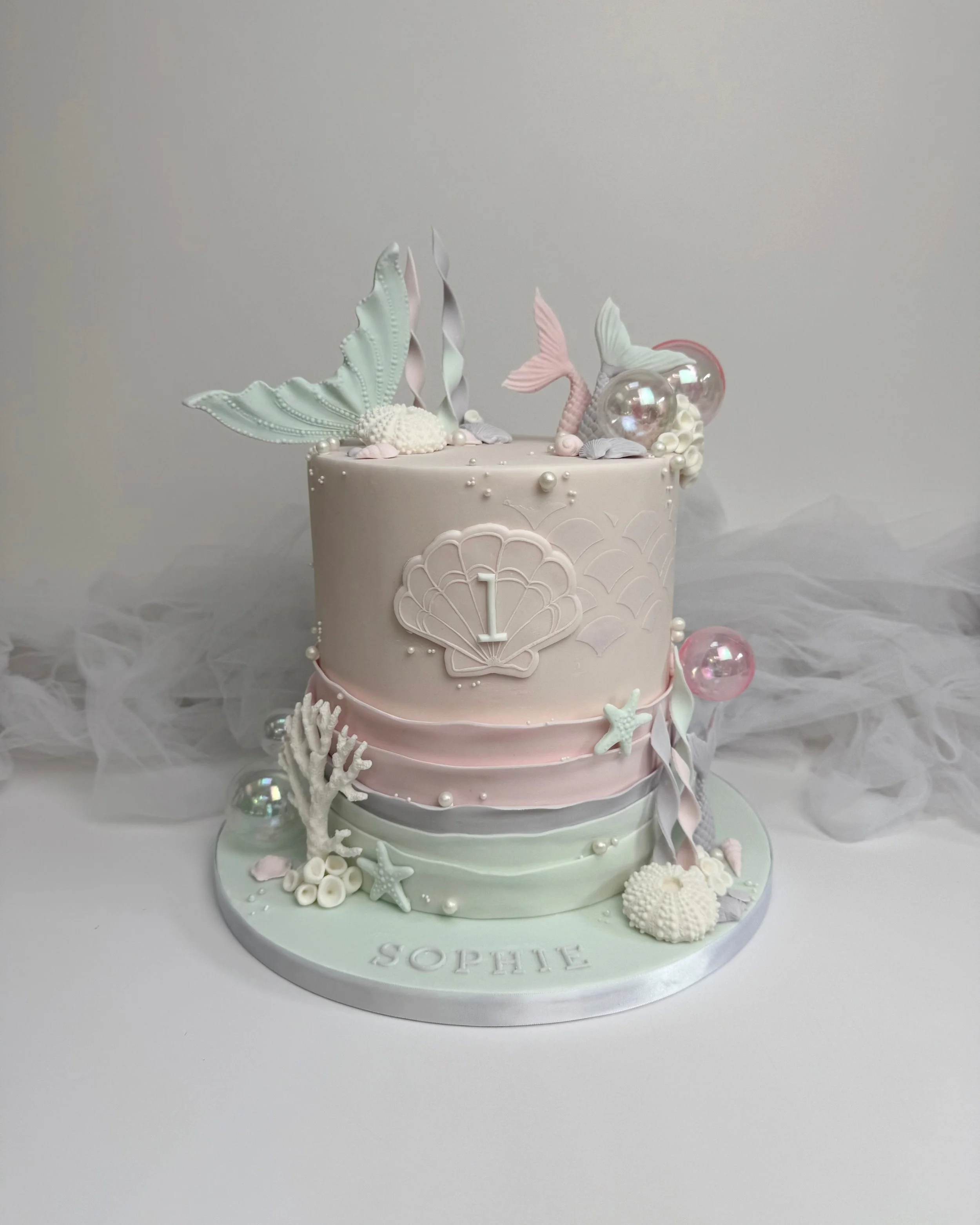 A two-tiered cake decorated with seashell, starfish, coral, and mermaid tail decorations, with a mermaid theme, including an embossed shell with the number '1' on the front, and the name 'Sophe' on the cake board.