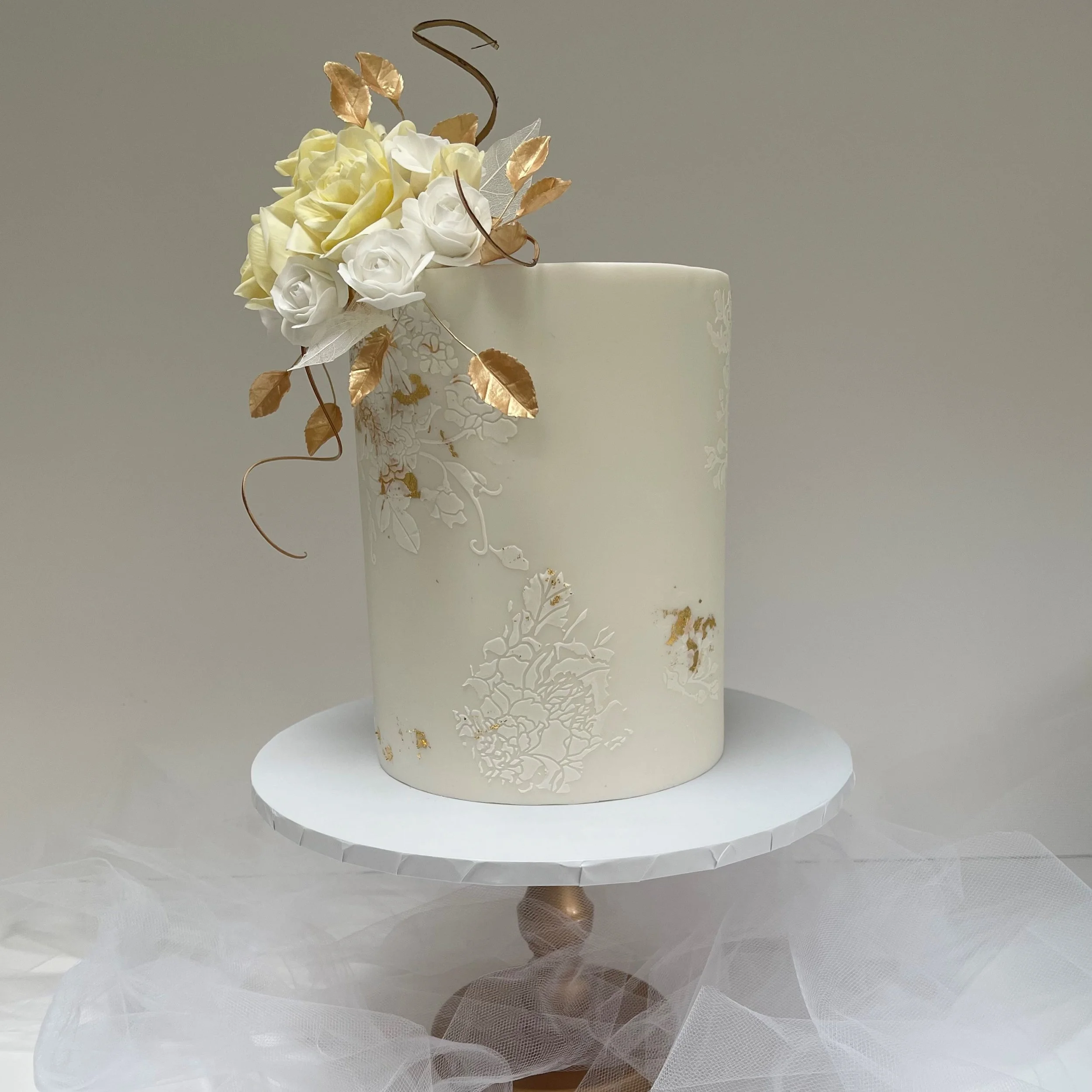 Elegant white wedding cake with floral decoration, gold accents, and a gold letter S topper, displayed on a cake stand with tulle fabric underneath.