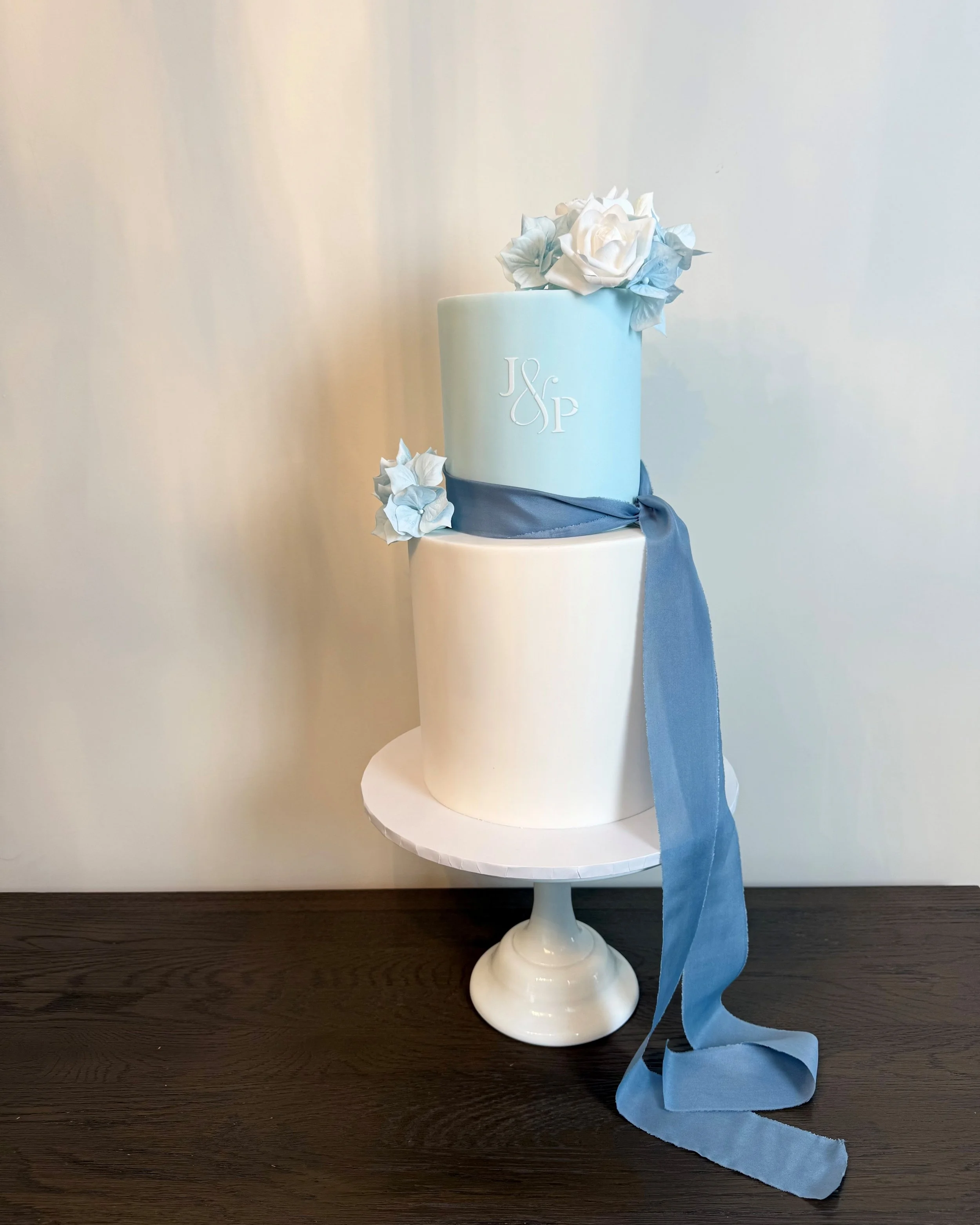 Two-tier wedding cake with white and light blue fondant, decorated with blue ribbons and white and blue flowers, monogrammed with J & P, on a white cake stand.