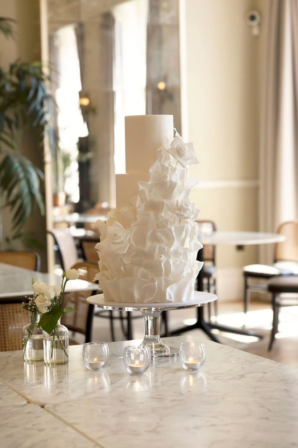 Elegant white wedding cake with ruffled design, placed on a cake stand with small candles and a flower arrangement on a marble table in a decorated reception area.