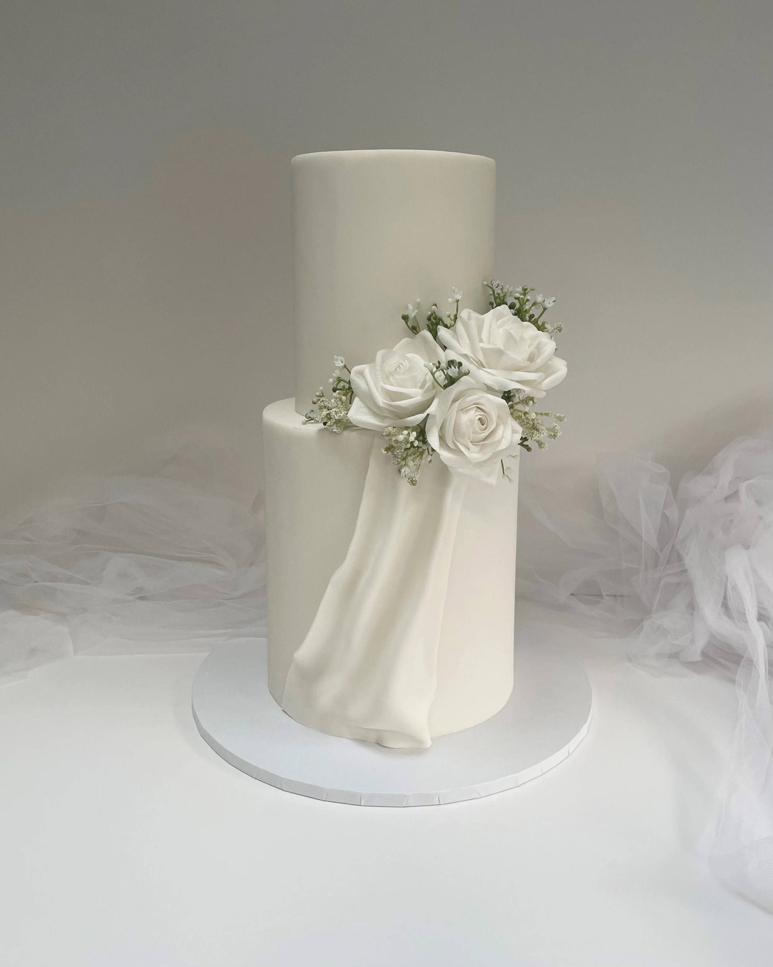 Elegant white wedding cake with two tiers, decorated with white roses and greenery, featuring a draped fondant detail on the lower tier, placed on a round white cake board.