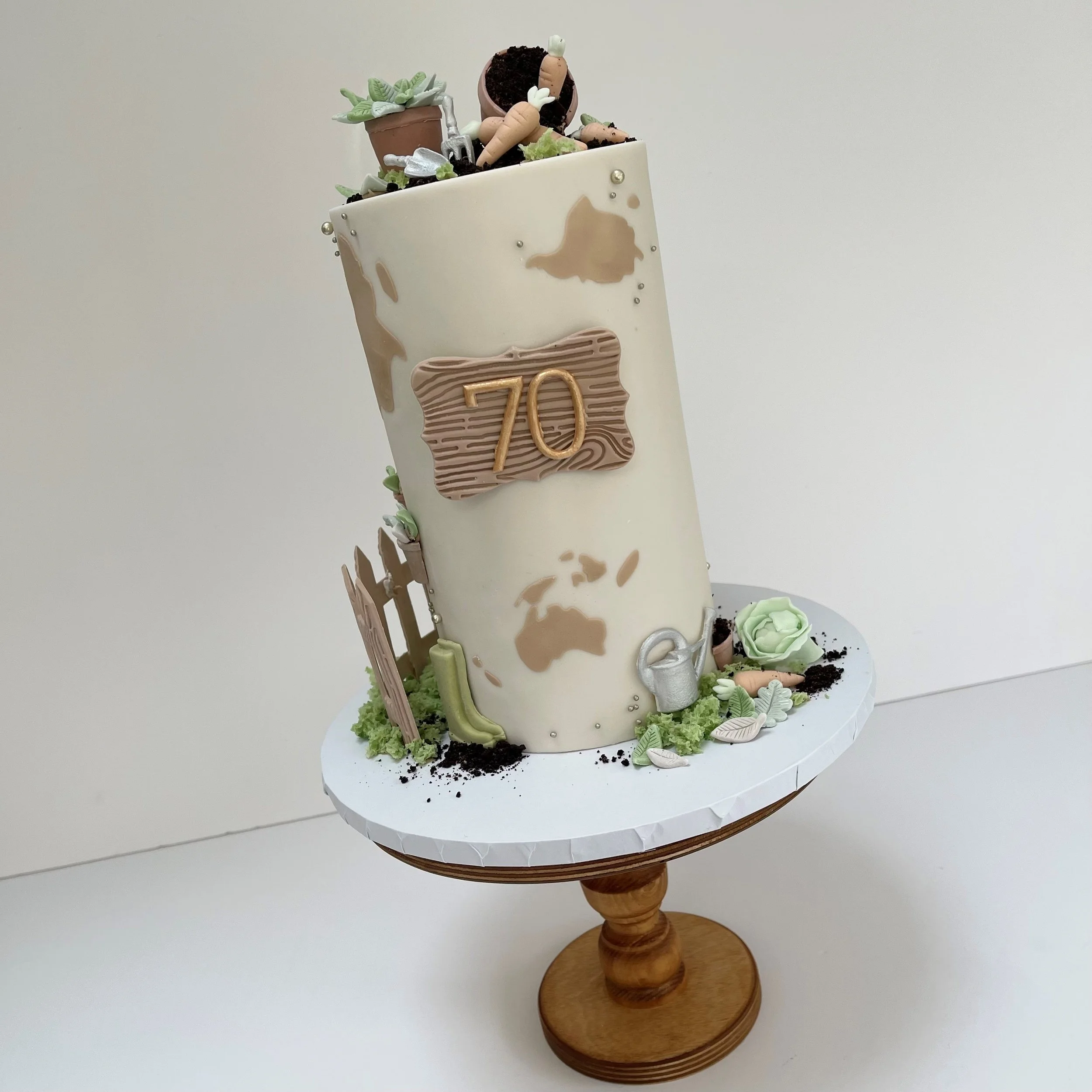 A tall cake decorated with gardening tools and plants, featuring a "70" anniversary plaque on the side, placed on a wooden cake stand.