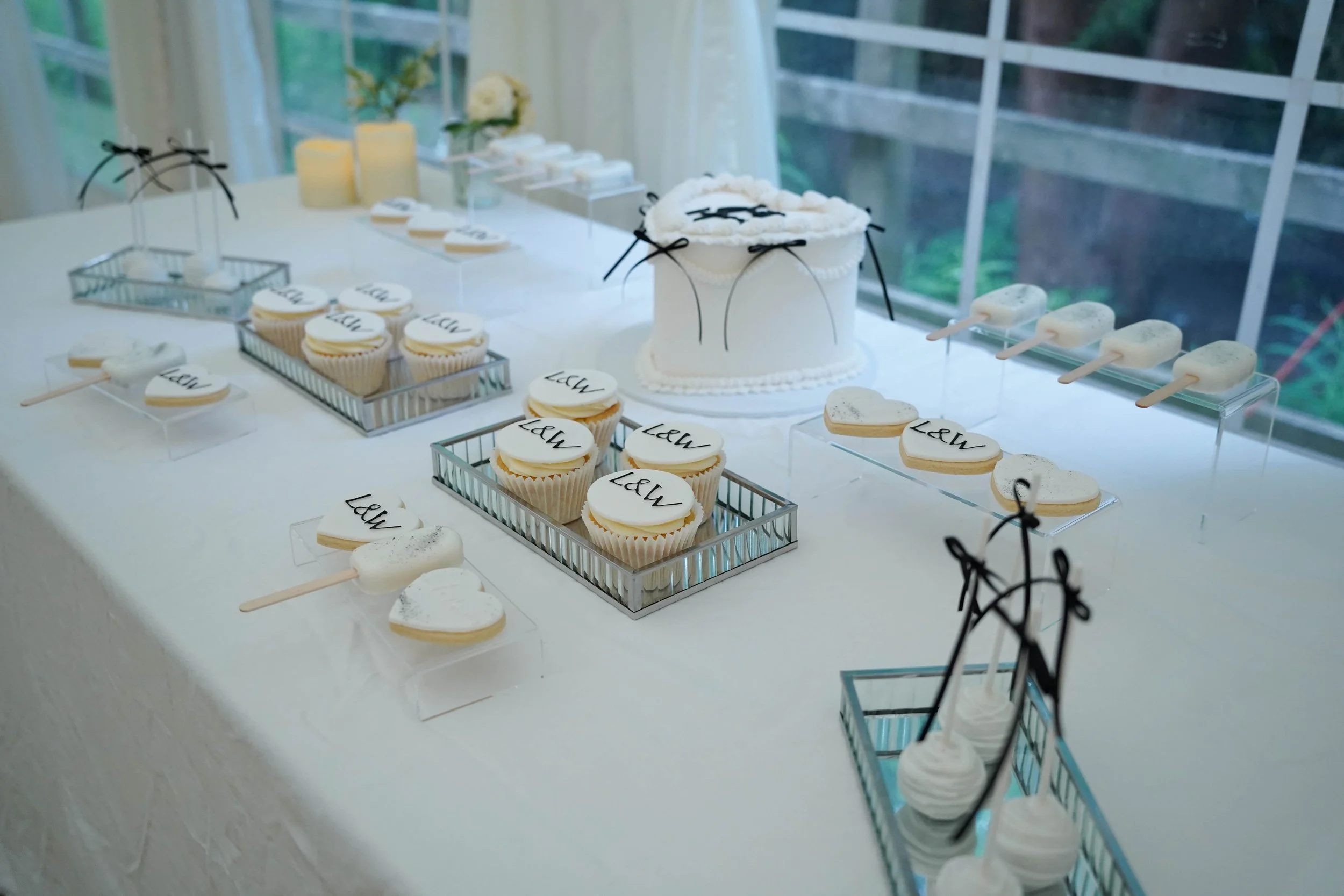 A table decorated for a celebration with white and black themed desserts, including cupcakes, cookies, and cake pops, with a white cake in the center and black ribbons, set near a window with a garden view.