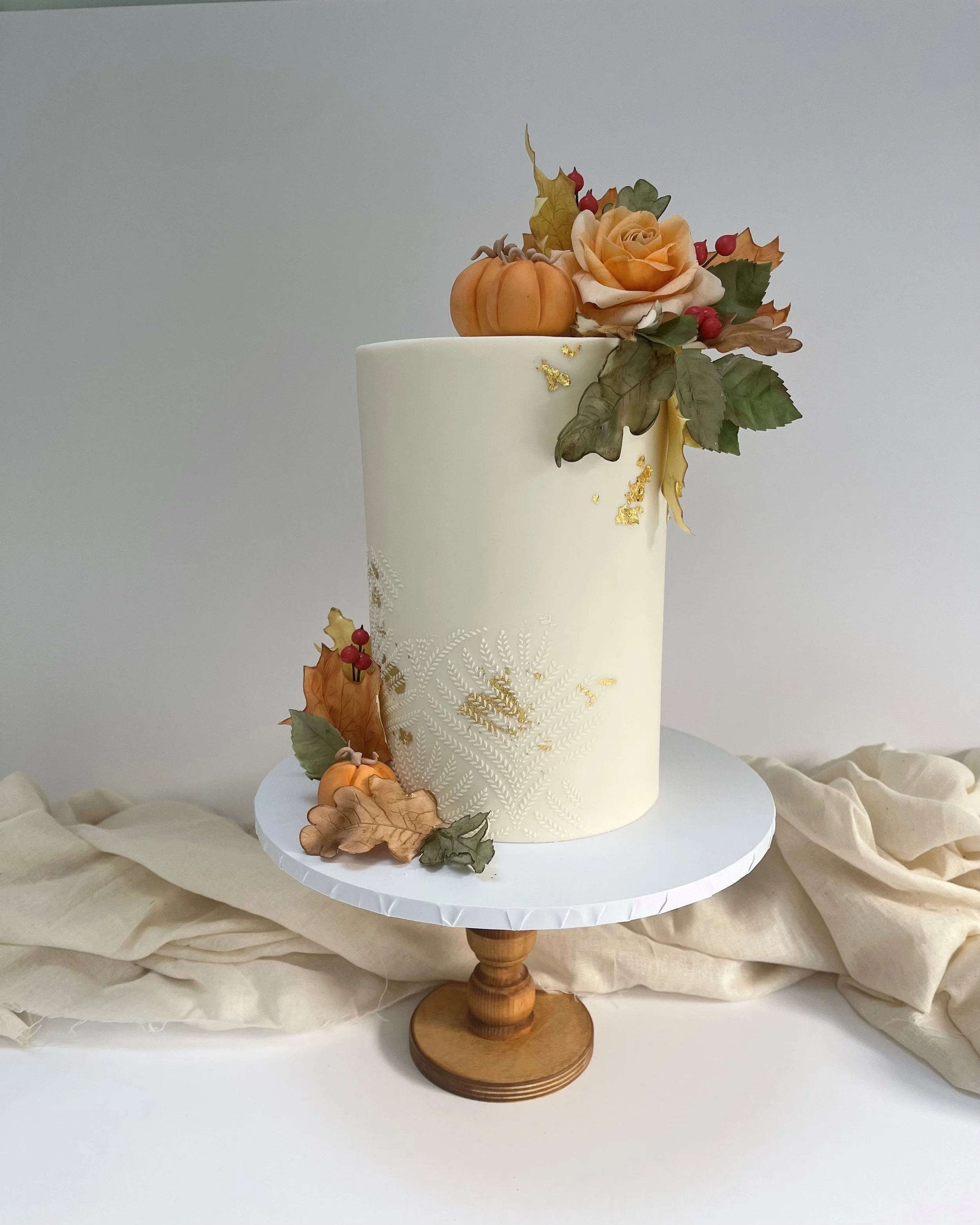 Elegant white cake decorated with autumn-themed flowers, leaves, small pumpkins, and gold accents, displayed on a wooden cake stand with a beige cloth backdrop.