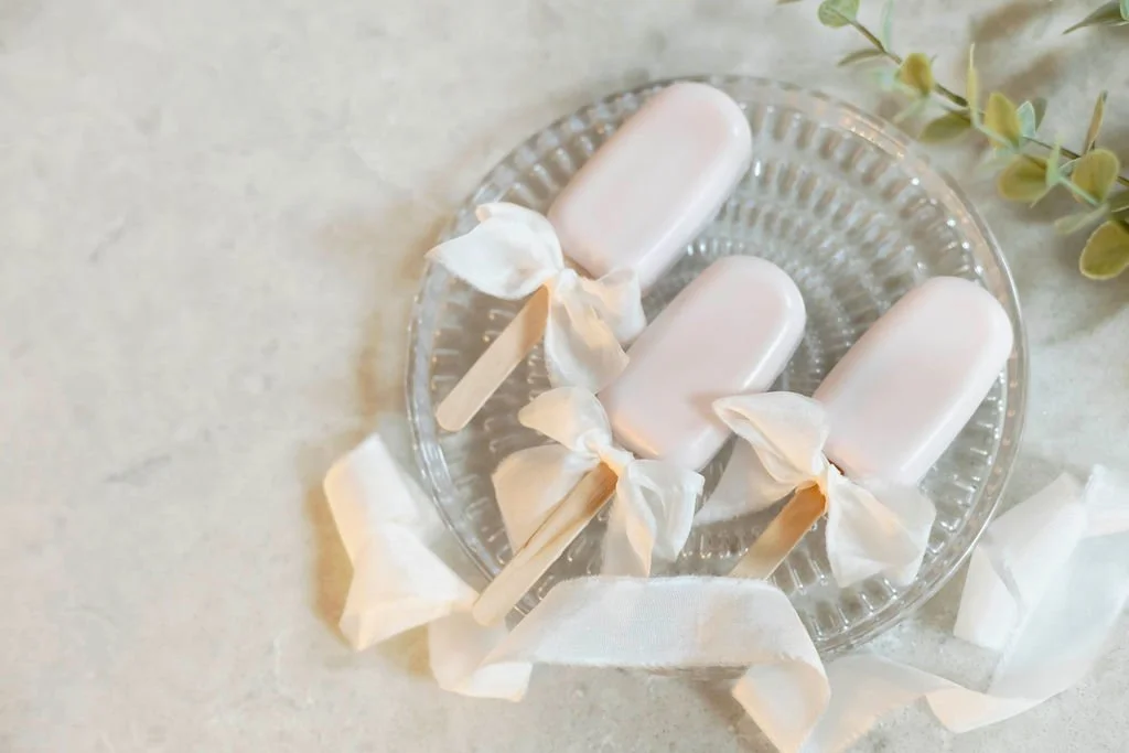 Three white popsicles wrapped with paper and tied with ribbons on a clear glass plate, with a sprig of greenery nearby.