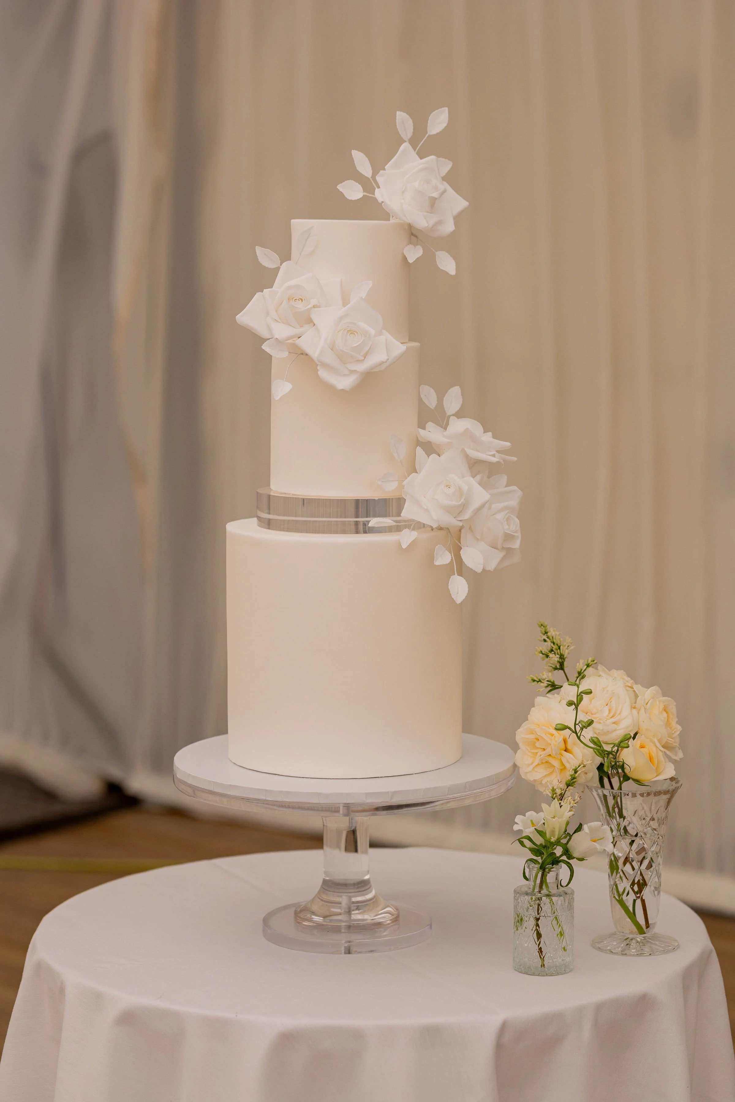 Elegant white three-tiered wedding cake decorated with white roses and leaves, placed on a glass cake stand, with additional flowers in vases on a table.