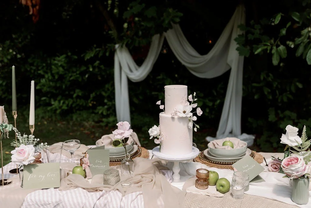Elegant outdoor table setting with a white cake decorated with flowers, surrounded by plates, glasses, and green apples, with a backdrop of greenery and draped white fabric.