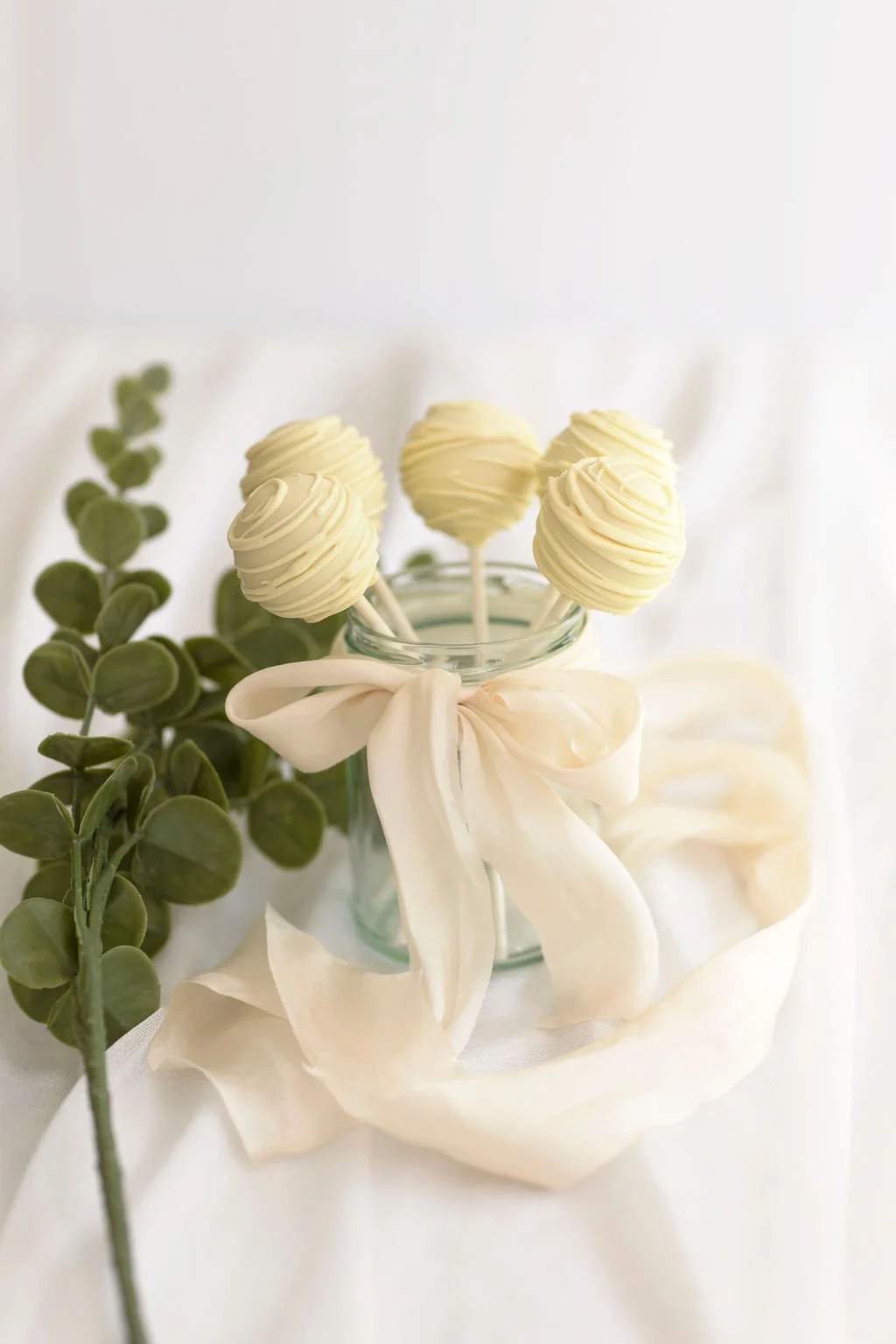 A glass jar with a cream-colored ribbon tied in a bow, containing five white chocolate cake pops decorated with swirled patterns on sticks, next to green foliage on white fabric.