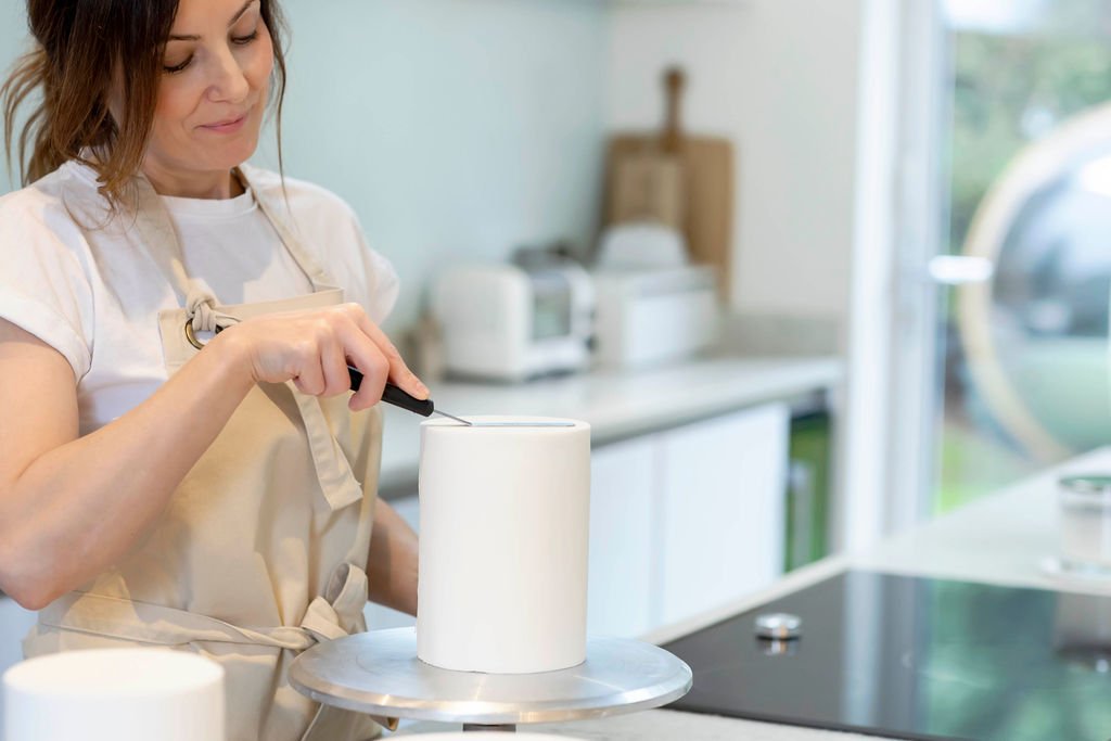 Rebecca is icing a wedding cake. She is wearing an apron over her white tshirt and has her brown hair tied back.