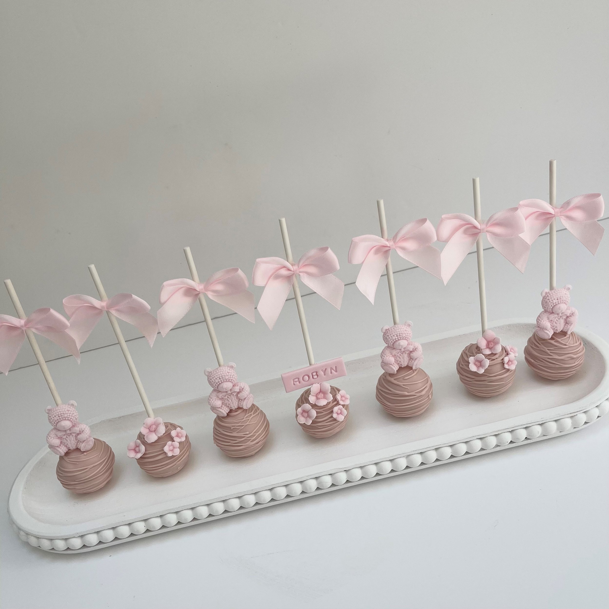 Decorative tray of pink cake pops with teddy bear and bow toppers, with a pink name tag reading 'Robyn'.