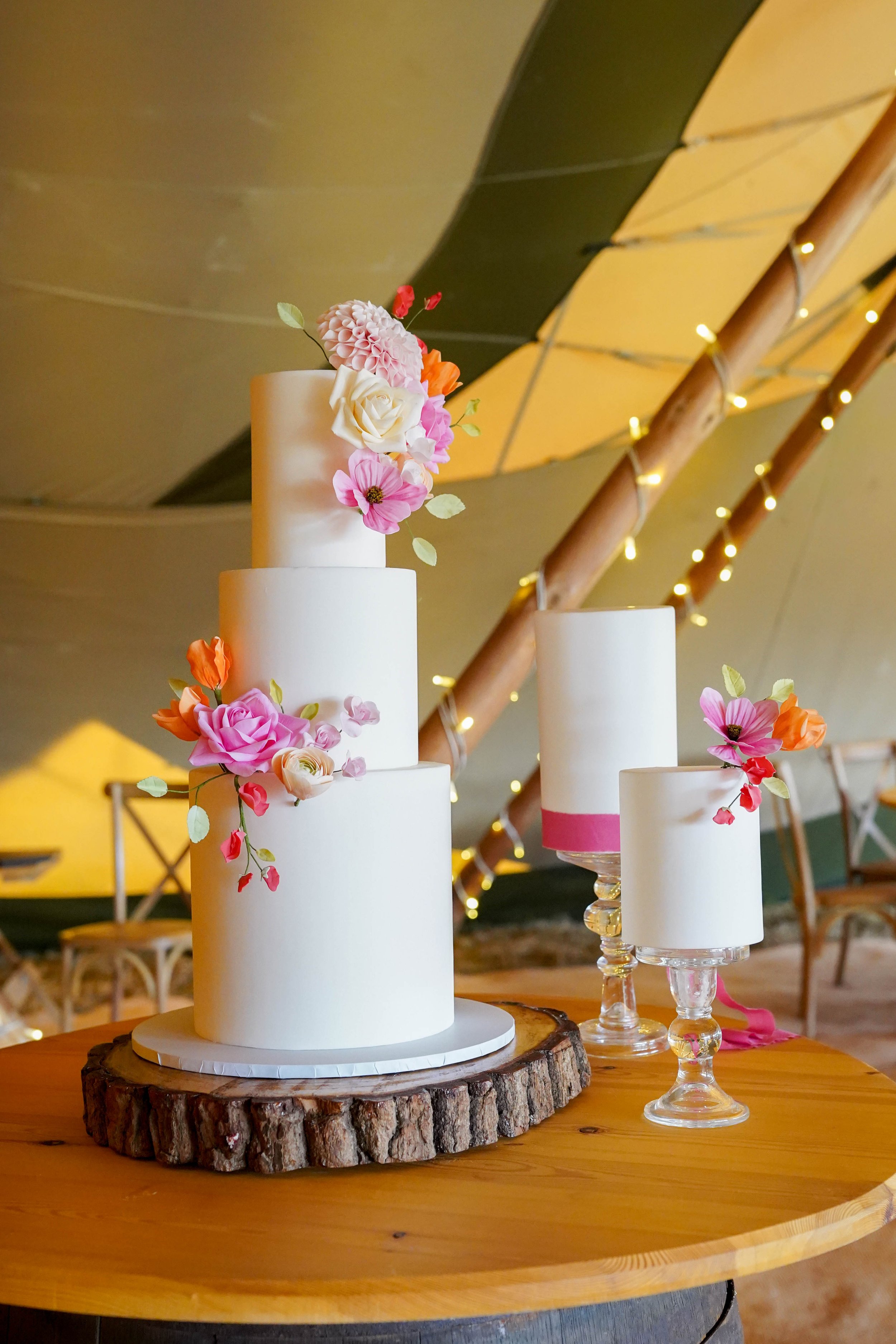 A set of three white wedding cakes decorated with pink, white, and orange flowers on a wooden table.