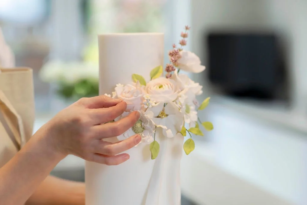 A person arranging a white floral cake with pink and white flowers and green leaves.