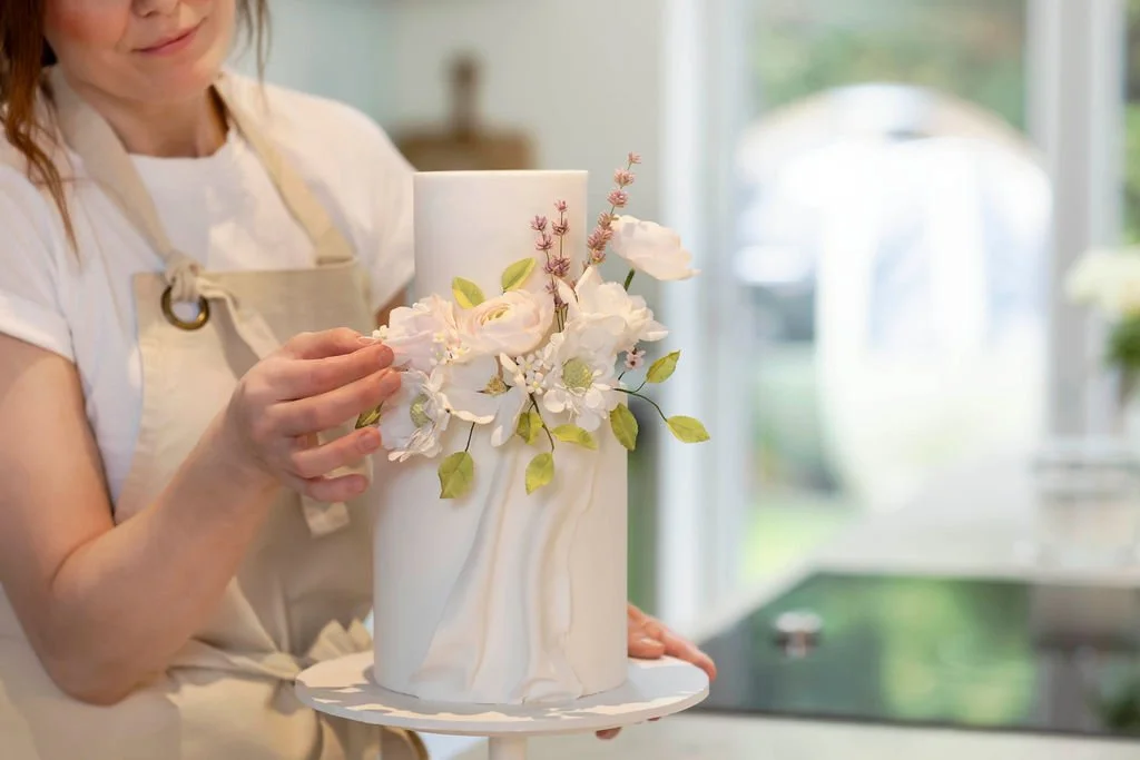 Rebecca is decorating a white cake with pink and white sugar flowers in a bright kitchen.