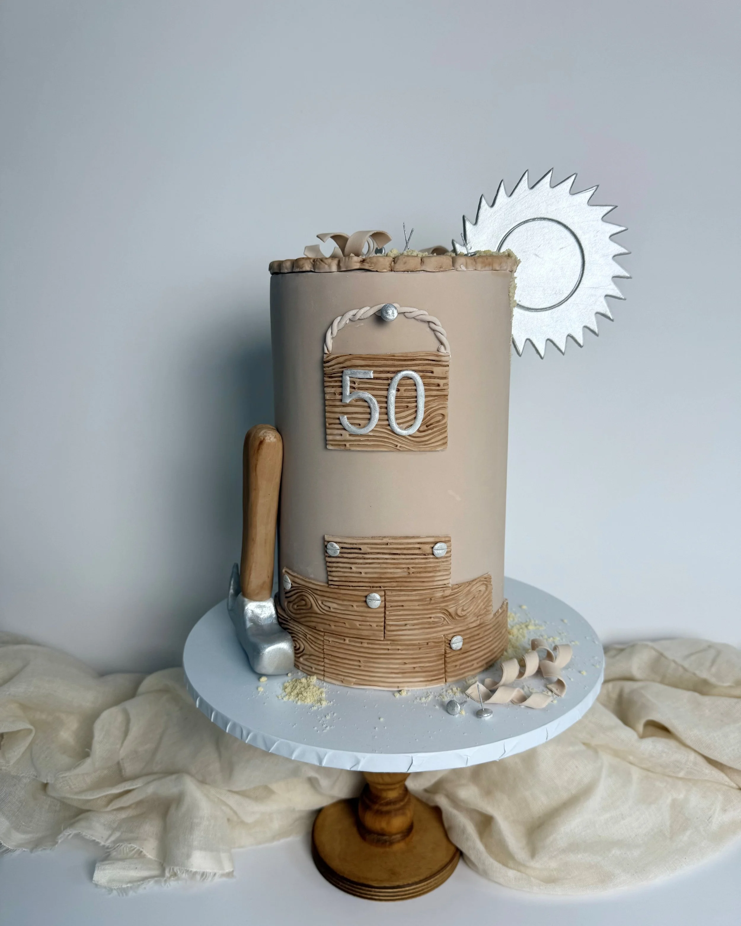 Cake decorated with a wooden theme, displaying the number 50 on a wooden sign, with a small hammer, wood shavings, and a sun-shaped cake topper.