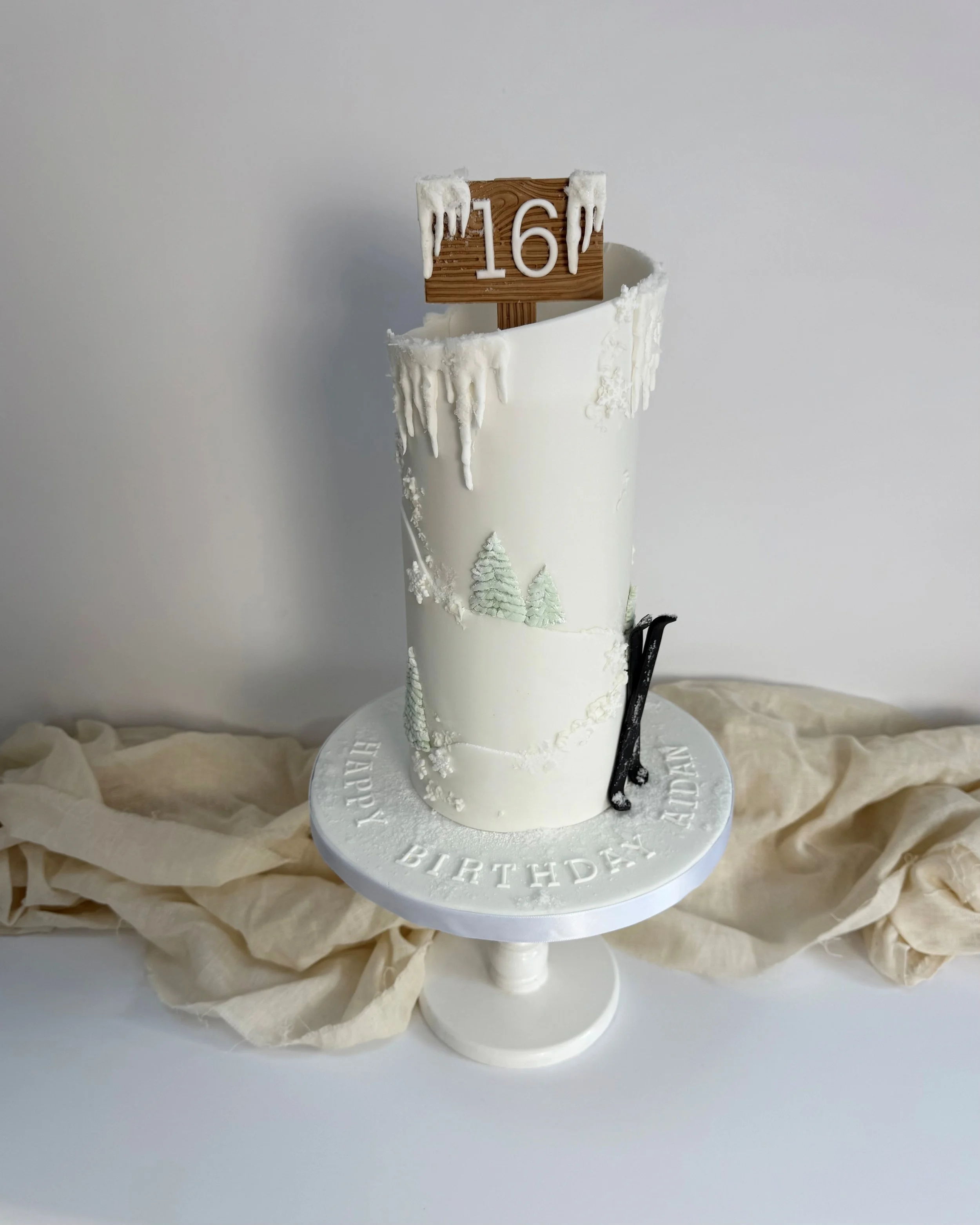 A winter-themed birthday cake with white icing, decorated with trees and snow-like details, featuring a wooden sign with the number '16' and a black and white hockey stick, on a white cake stand with 'Happy Birthday' engraved around the edge, sitting