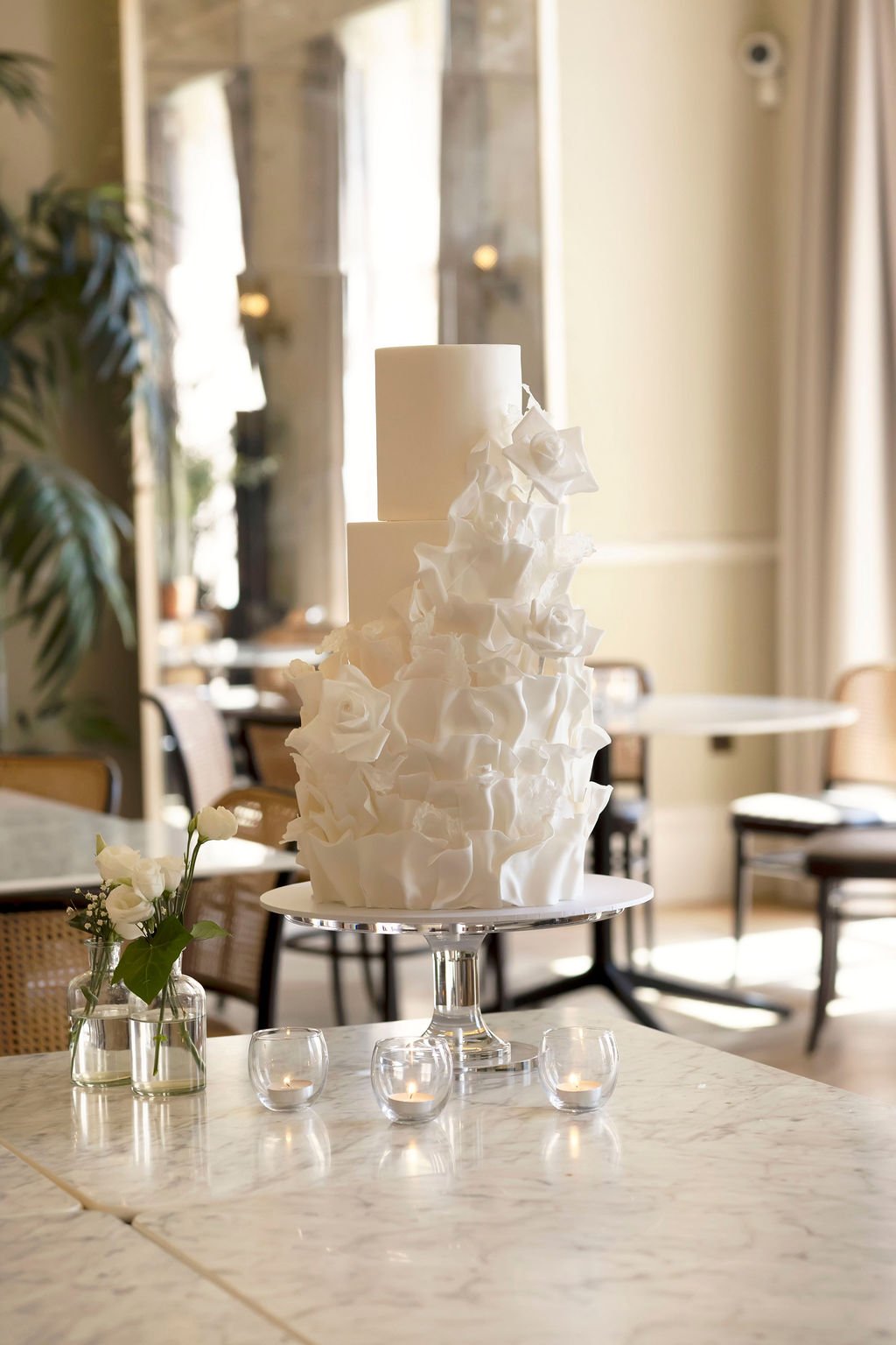 A tall white wedding cake with a ruffled design on a white cake stand, on a marble table decorated with white flowers in a glass vase and small candles, in a well-lit room.