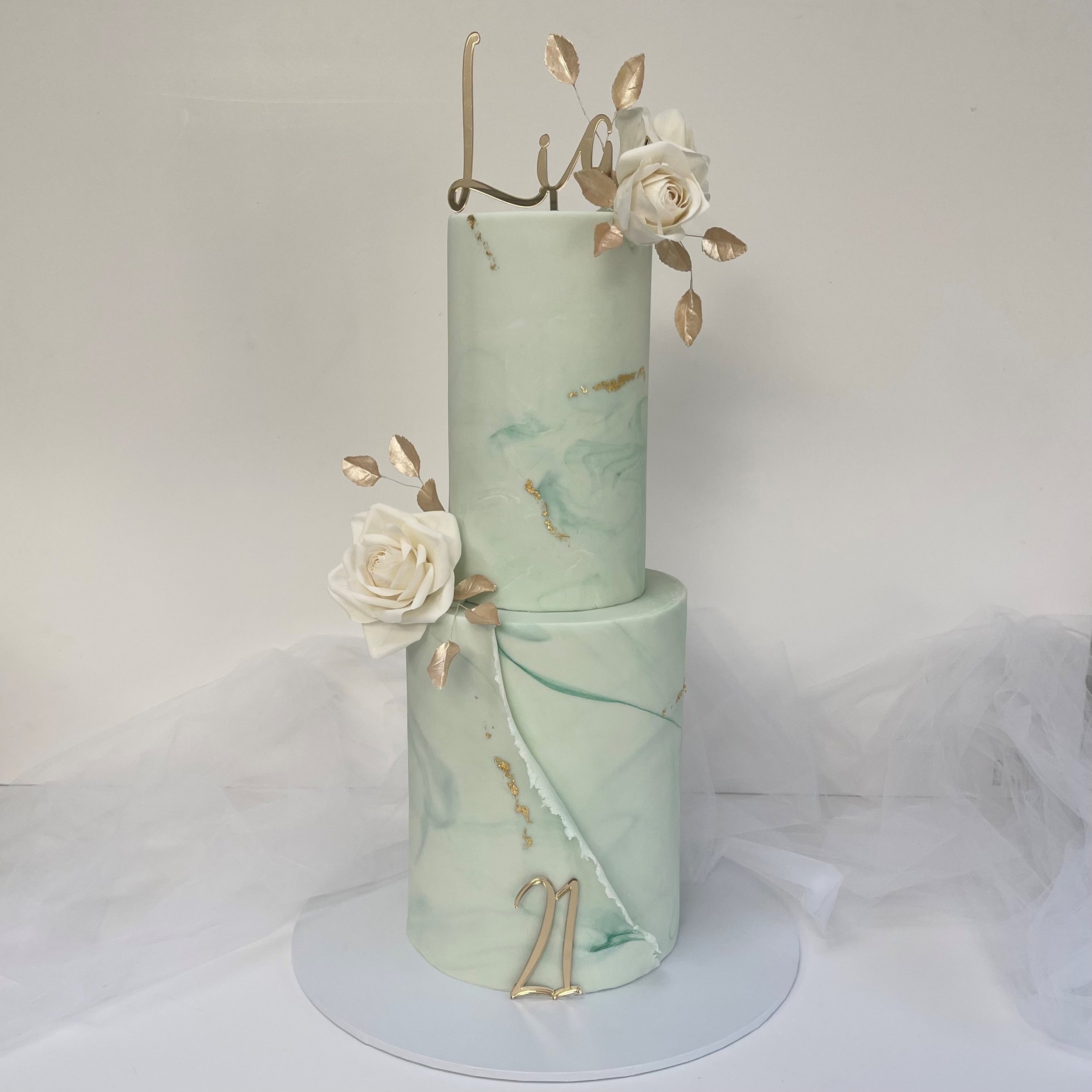 Two-tiered wedding cake with a green marble pattern, decorated with white roses, gold leaves, a gold '21' topper, and a gold 'Lia' cake topper.