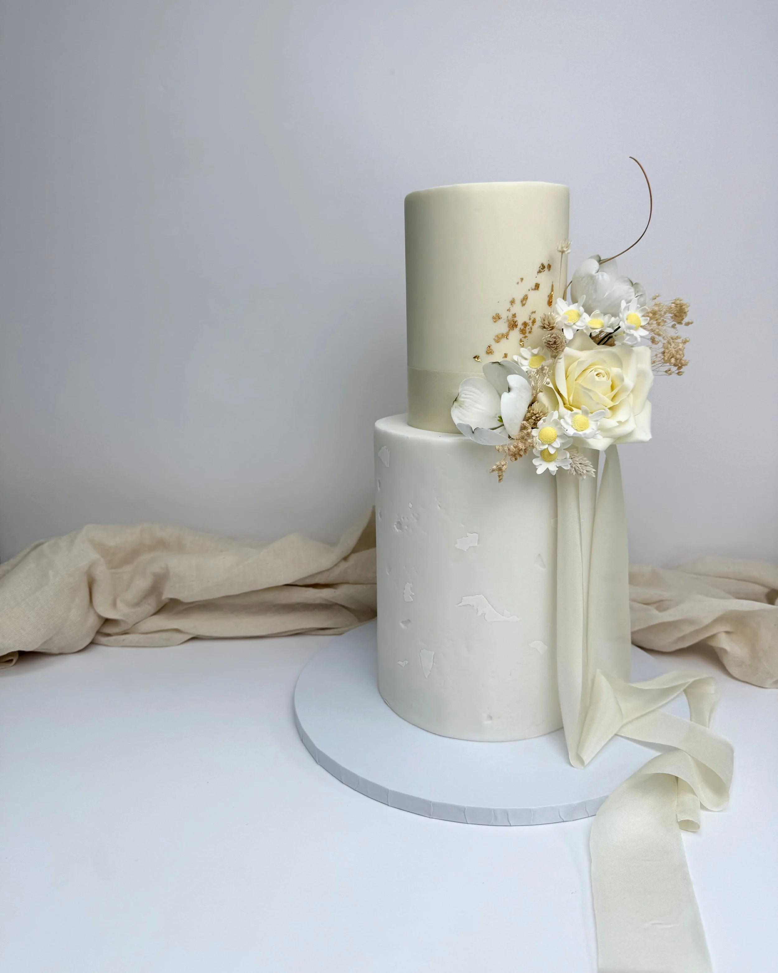 Two-tiered white wedding cake decorated with white and cream-colored flowers and a beige ribbon, placed on a white platform with beige fabric in the background.