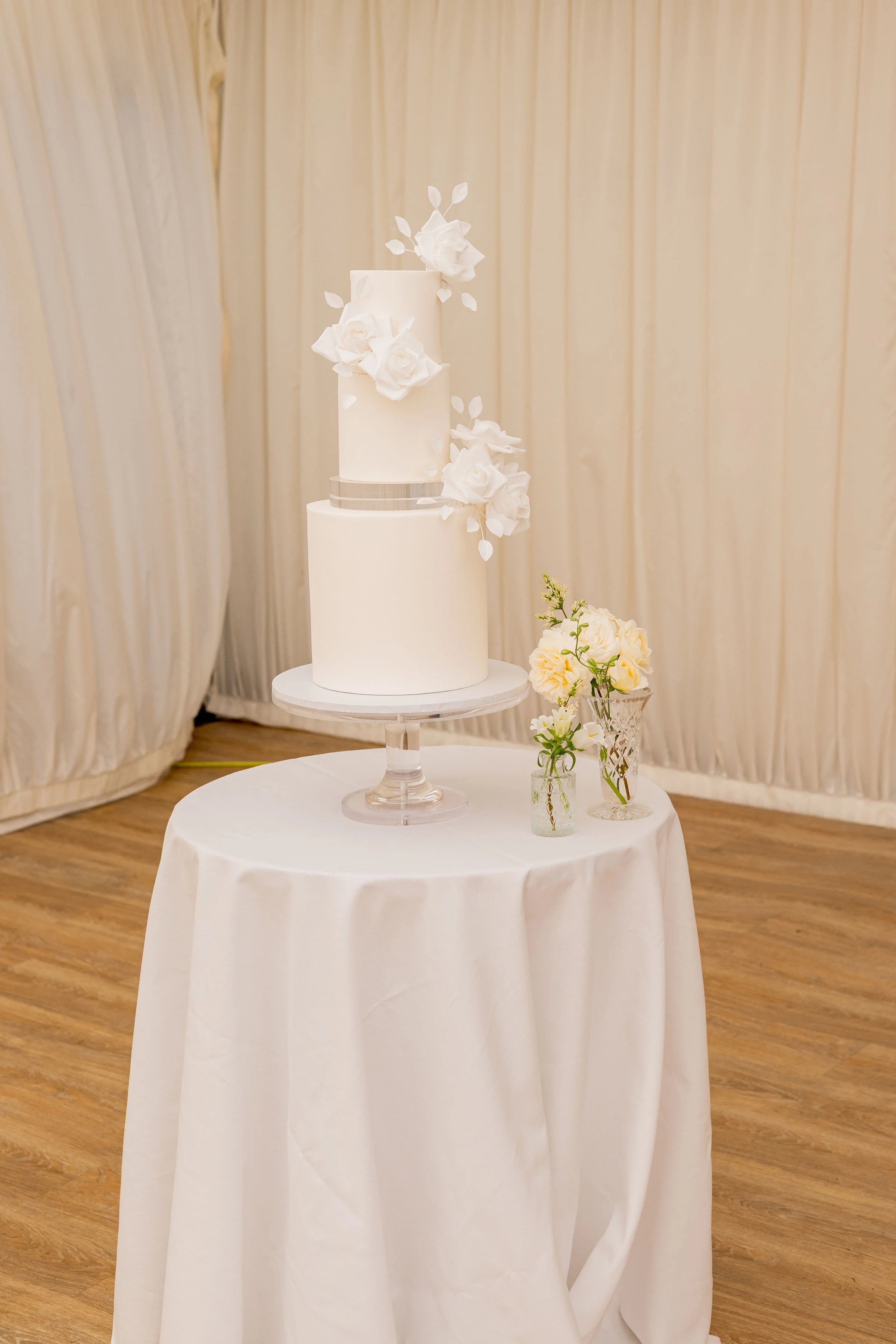 Elegant white two-tier wedding cake with floral decorations, displayed on a glass cake stand on a round white table with small flower arrangements in glass vases.