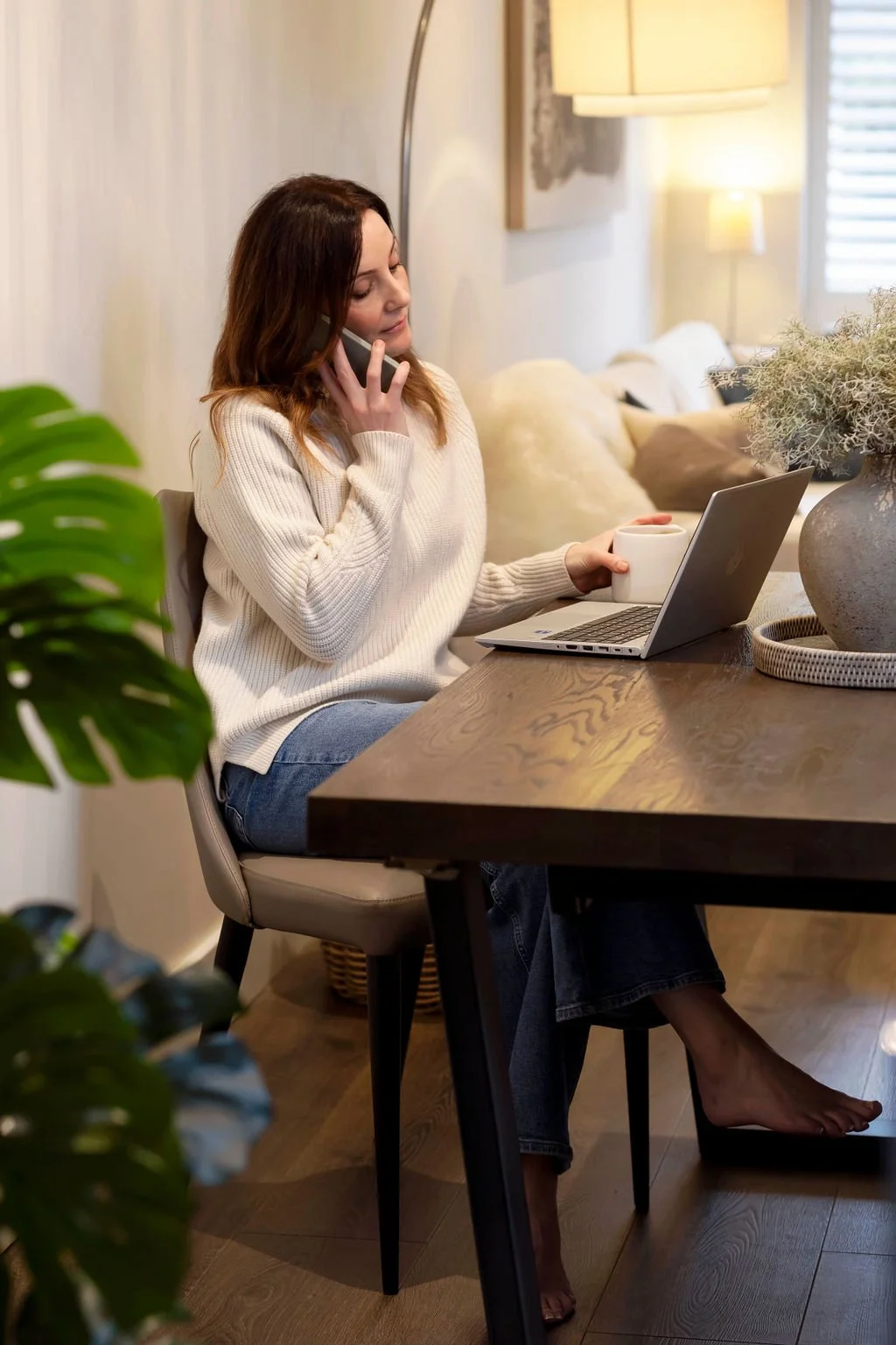 Rebecca is sitting at a dining table with a laptop, talking on the phone, holding a mug, in a cozy living room with soft lighting and plants.