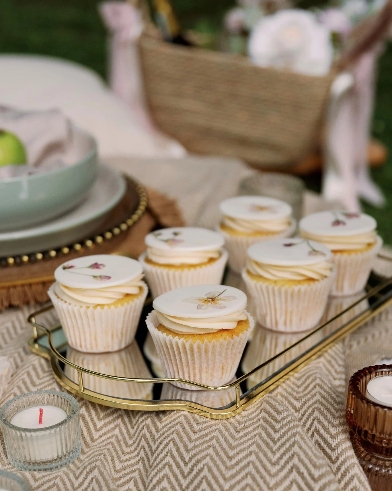 Cupcakes with white frosting and floral edible toppers on a gold wire tray, candles, and a wicker basket in the background, on a beige chevron tablecloth for a celebration.