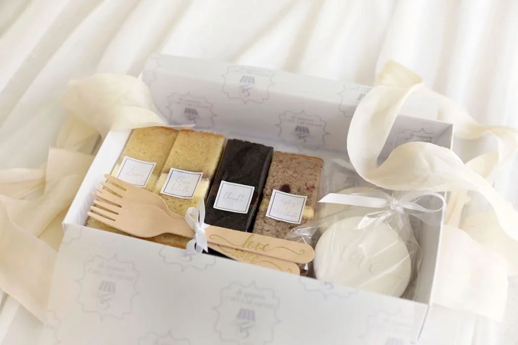 A gift box with four slices of cake, a small wooden fork, and a white wrapped item, set on a white surface with cream-colored ribbons.