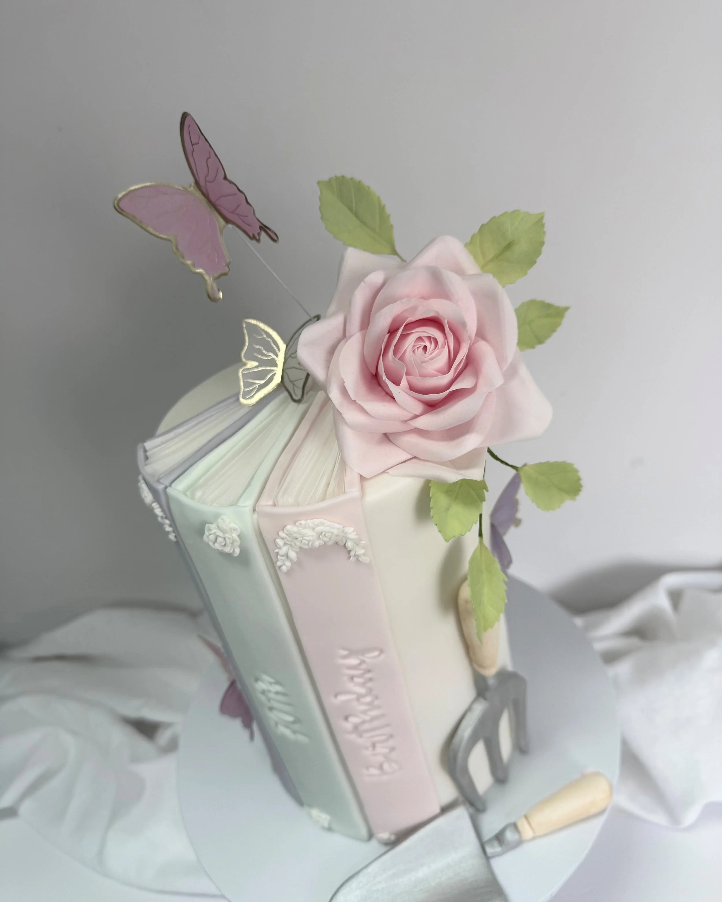 A cake designed to look like an open book with a pink rose, green leaves, butterflies, and gardening tools on it.