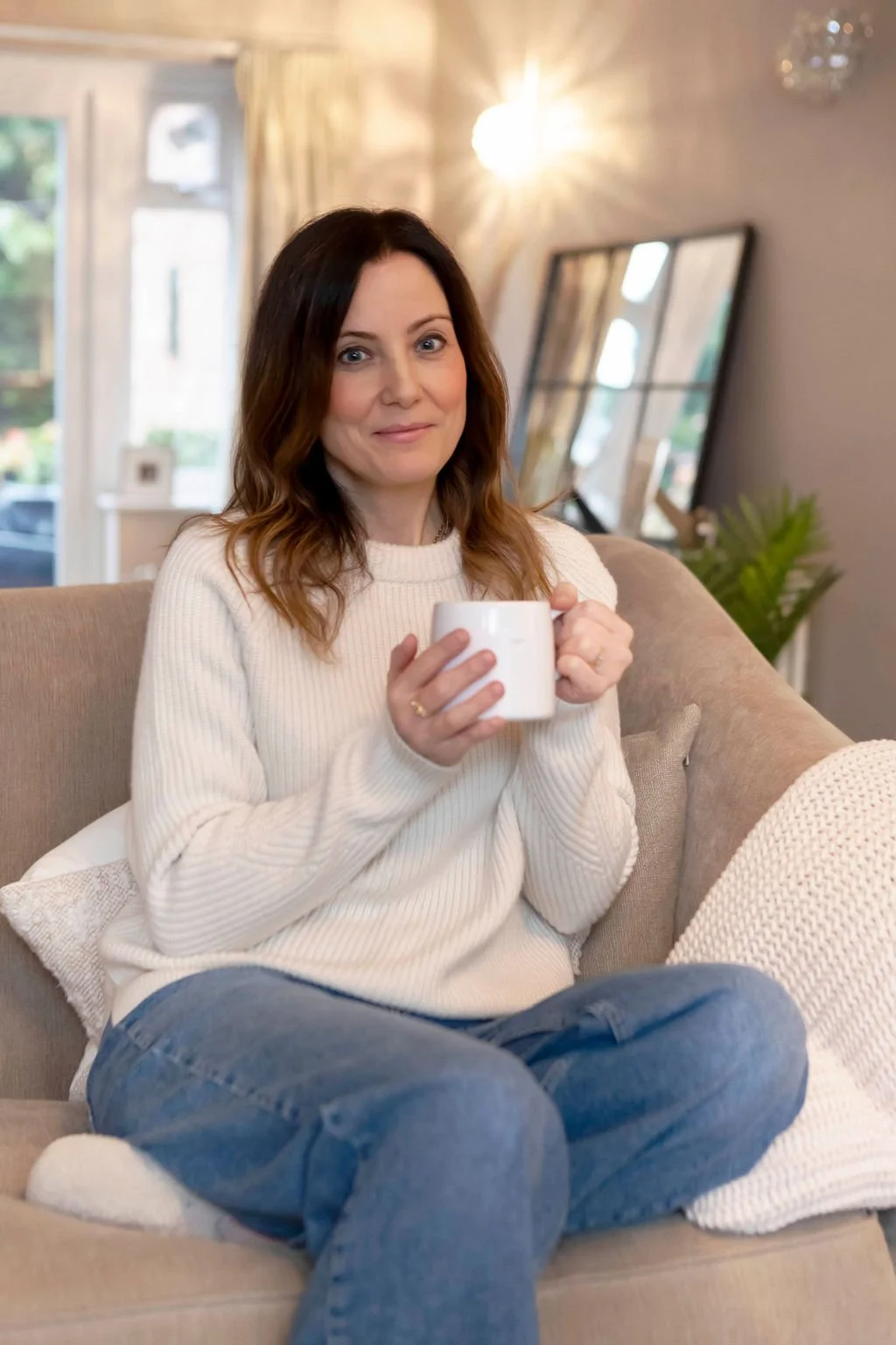 Rebecca is sitting on a beige sofa in a cozy living room, holding a white mug with both hands, wearing a cream-colored sweater and blue jeans, smiling softly at the camera.
