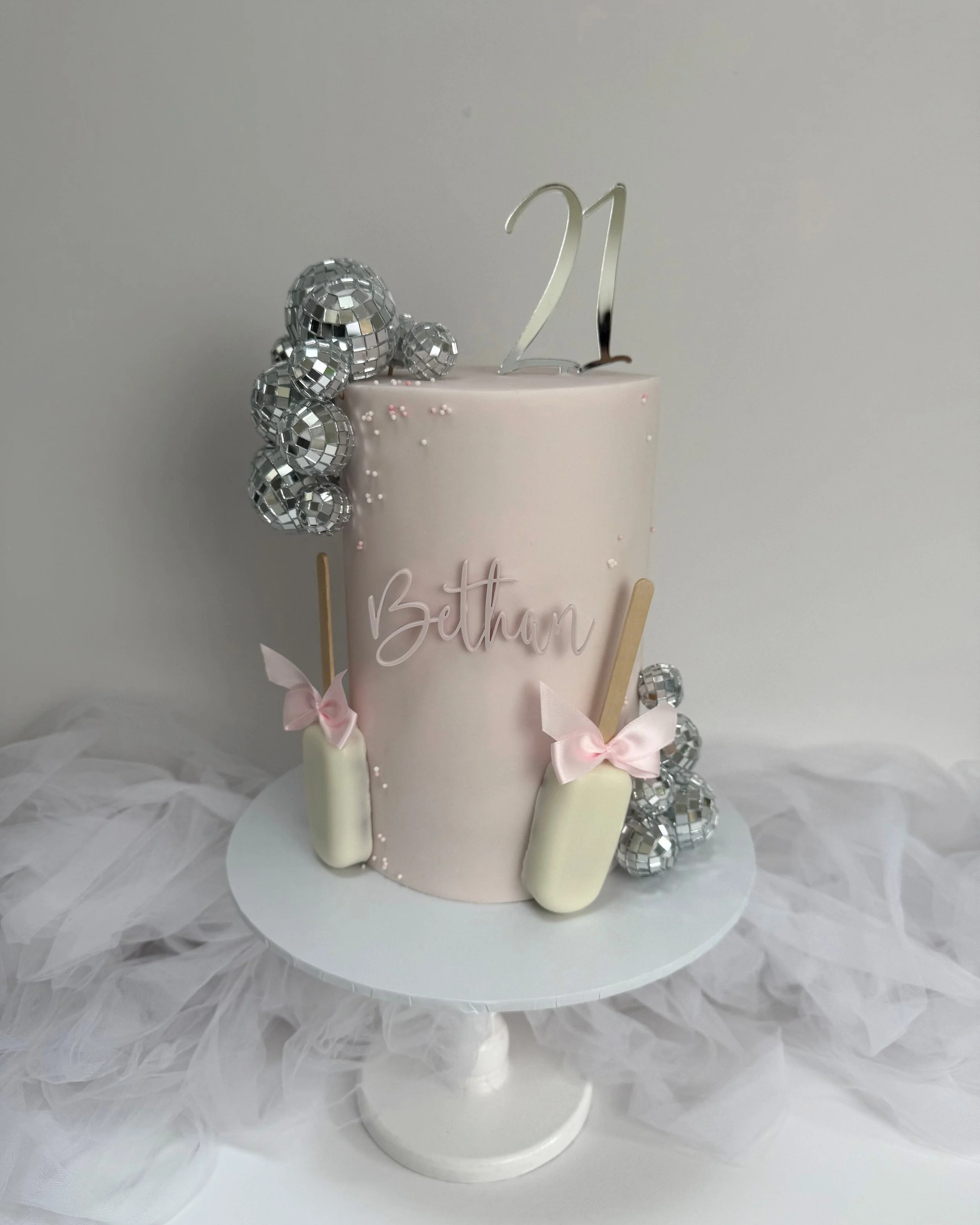 A pink birthday cake with silver disco balls, a silver '21' topper, and the name 'Bethan' written on it. The cake is decorated with pink bows and white popsicles and is displayed on a white cake stand with tulle fabric underneath.