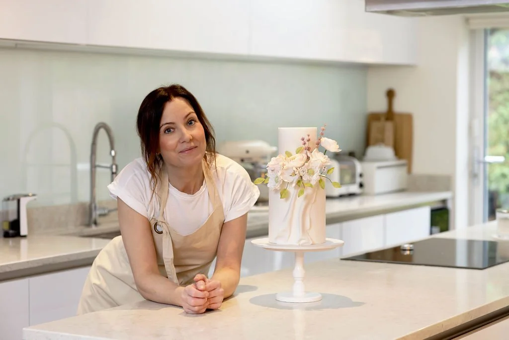 Rebecca is wearing a white t-shirt and beige apron leaning on a kitchen counter with a decorated white wedding cake with flowers on top.