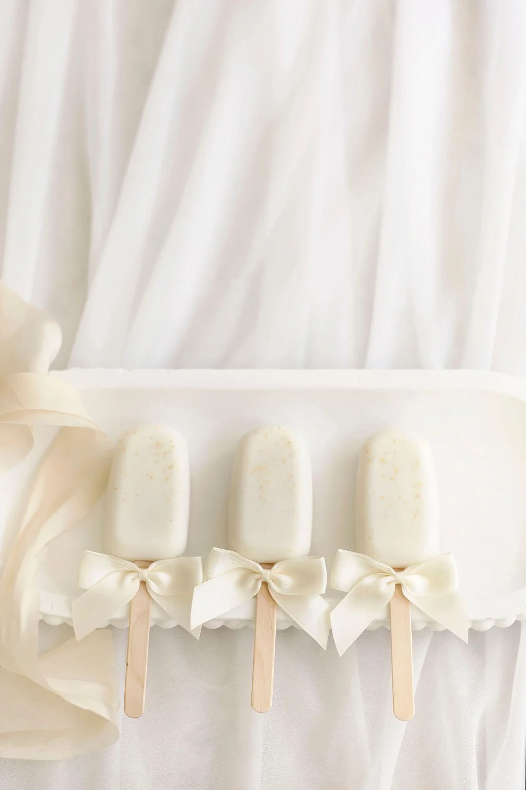 Three white popsicles with bows on sticks, placed on a white surface with white fabric in the background.