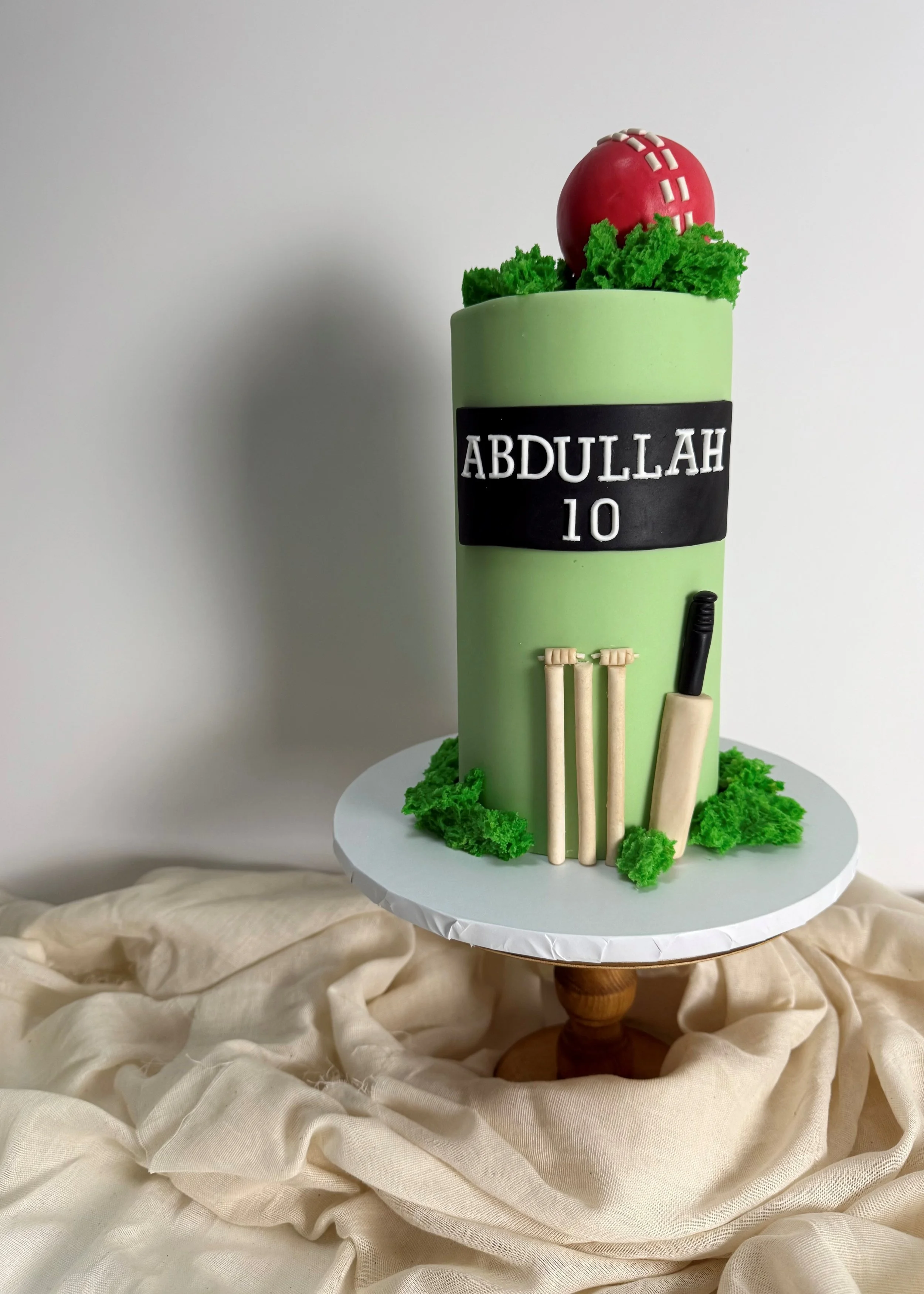A cricket-themed cake with green fondant, cricket bats, stumps, a ball, and a black scoreboard with the name 'Abdullah' and the number '10' on it, placed on a white cake stand.