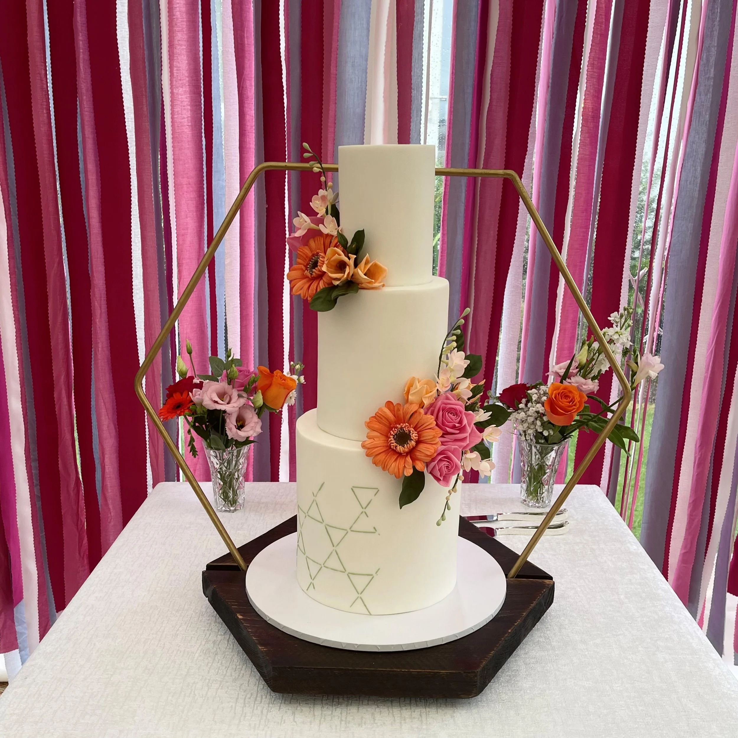 A multi-tiered white wedding cake decorated with pink and orange flowers, displayed on a wooden stand with a geometric frame, surrounded by floral arrangements on a table with pink and purple striped curtains in the background.