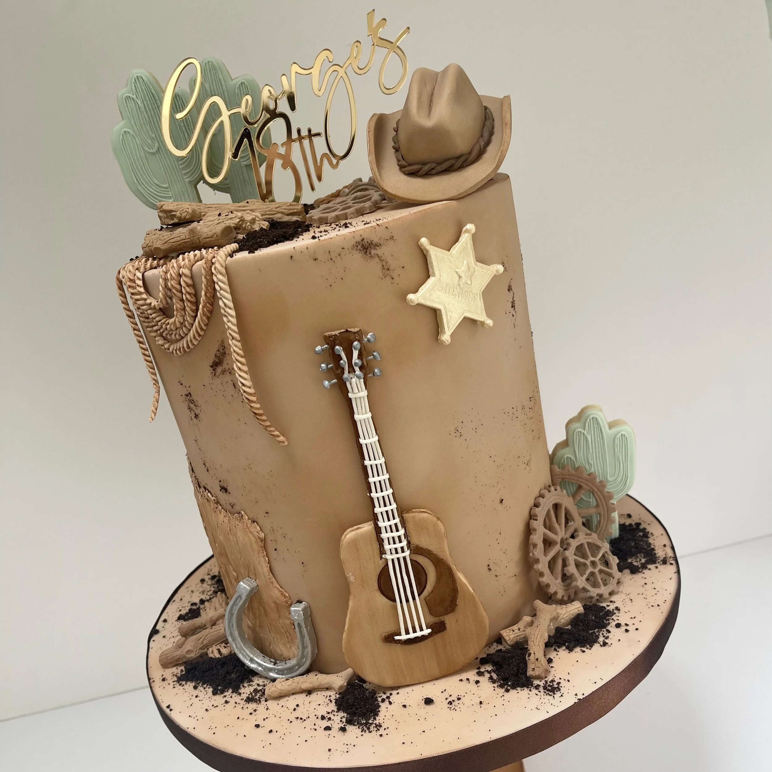 A cowboy-themed cake decorated with a gold 'Happy Birthday' topper, a guitar cake decoration, sheriff badge, horseshoe, cacti, gears, and desert-themed elements.